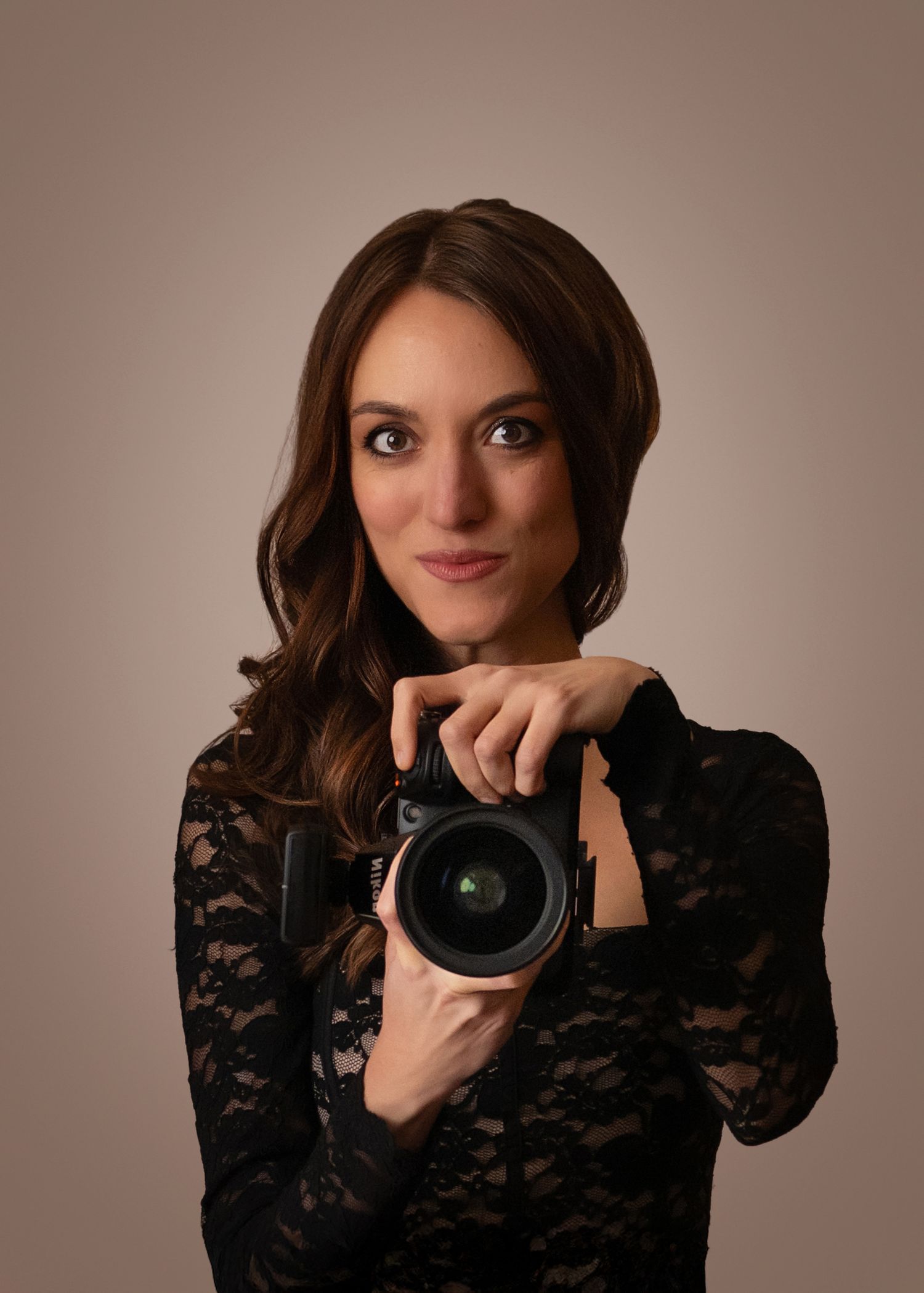A young brunette woman in black lace top holds a DSLR camera, posing confidently against a warm beige background.