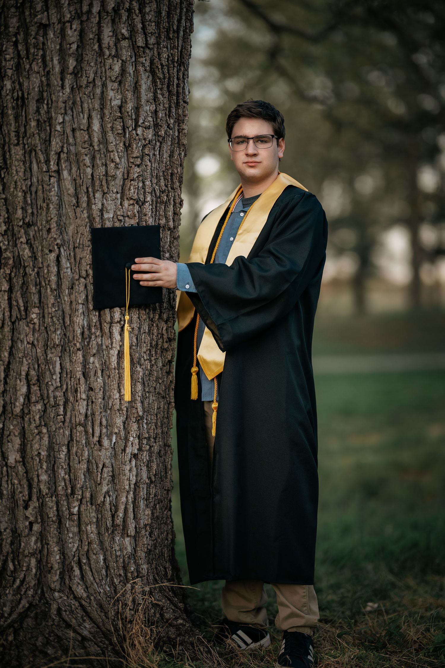 Young man in black and gold graduation gown and cap leans against a tree outdoors, smiling for senior photos.