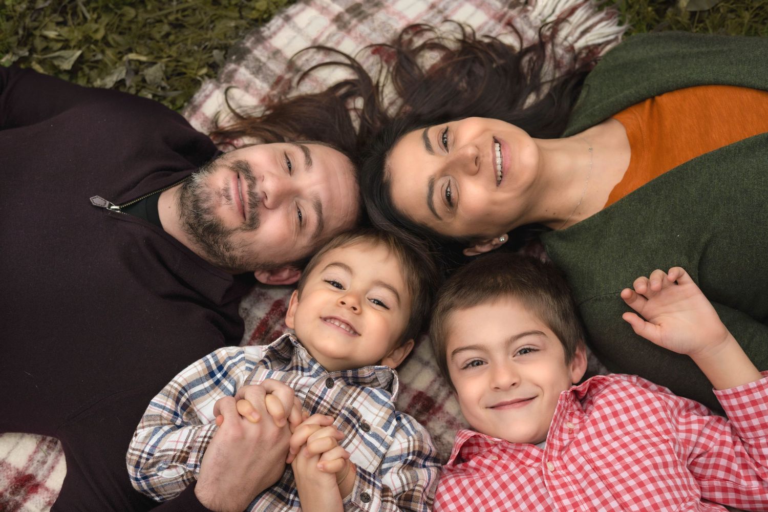 Family of four lying on a blanket outdoors, smiling up at camera, parents with two young children.