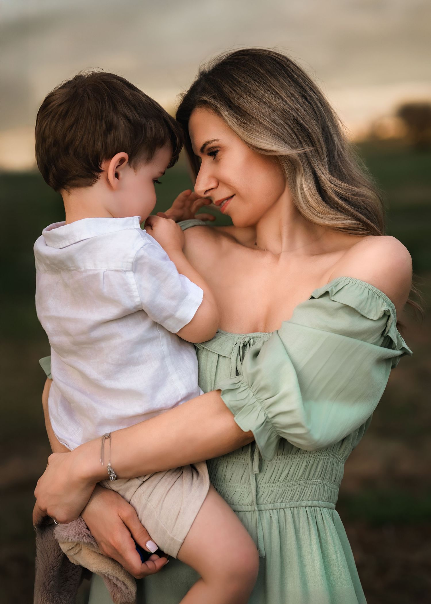 A mother in a sage green off-shoulder dress tenderly holds her toddler son outdoors at golden hour.