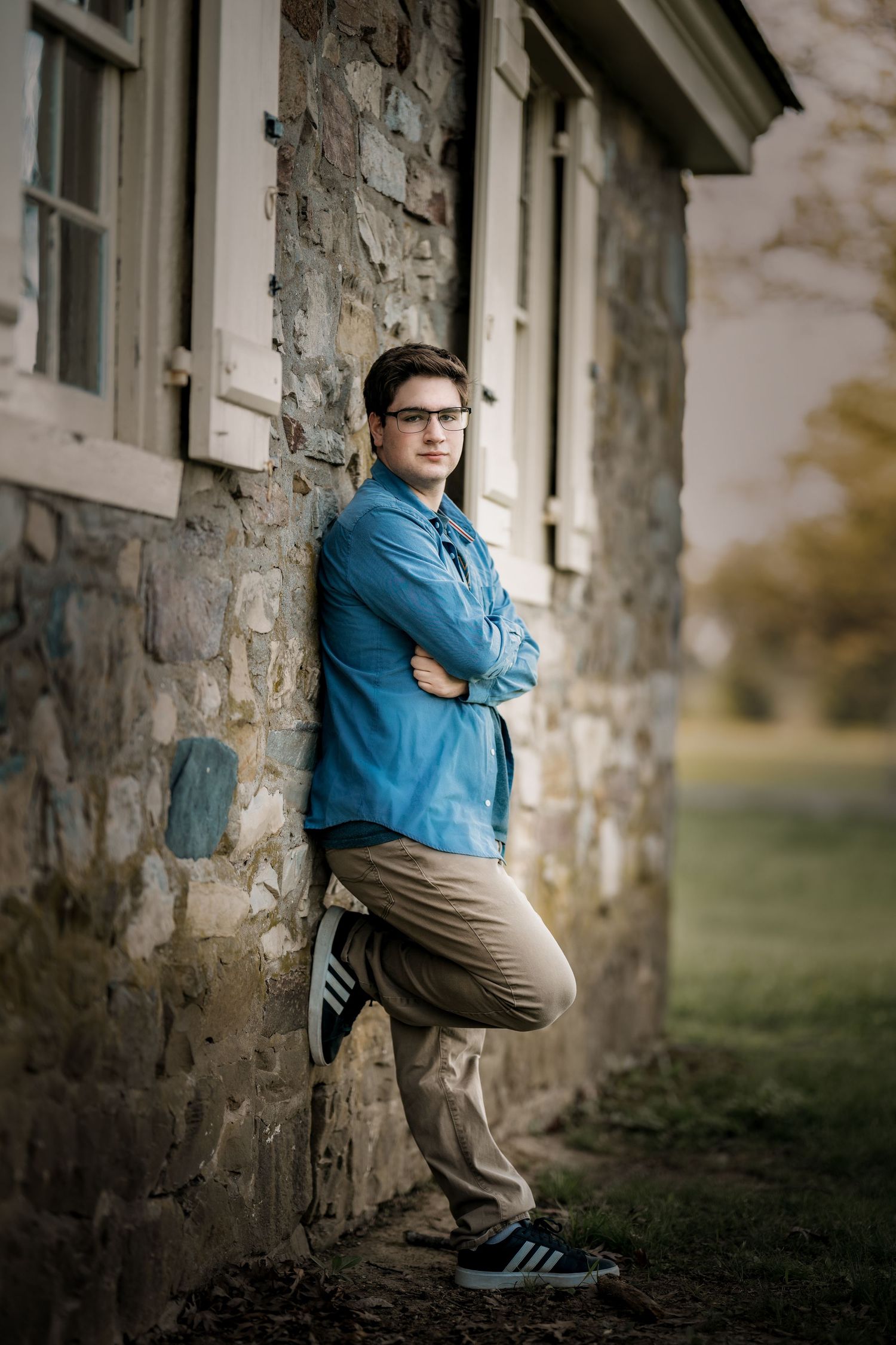 Young man in blue shirt and khakis leans against stone building wall with arms crossed wearing Adidas sneakers.
