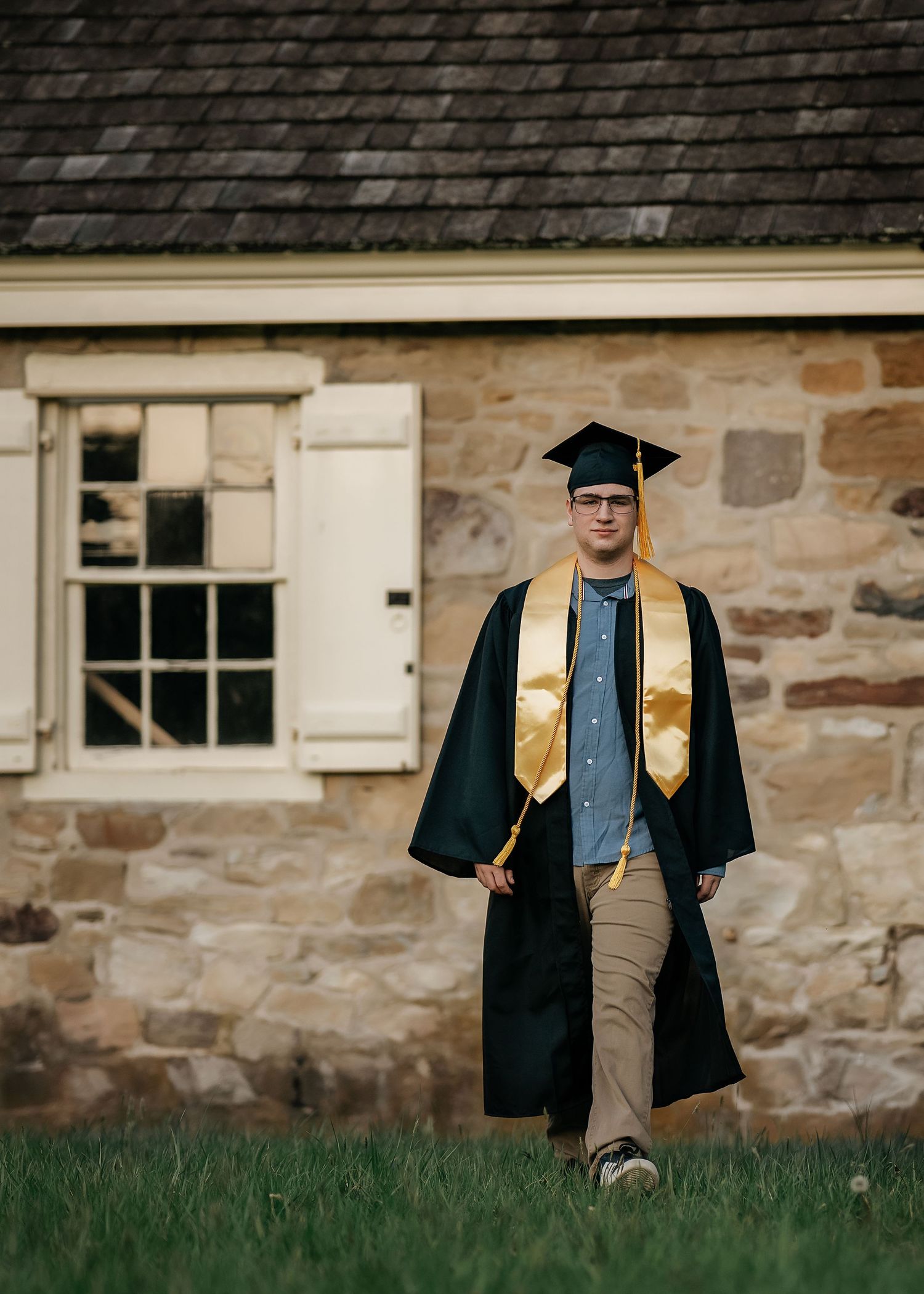 A young man in a dark green graduation gown and gold stole walks proudly in front of a historic stone building.