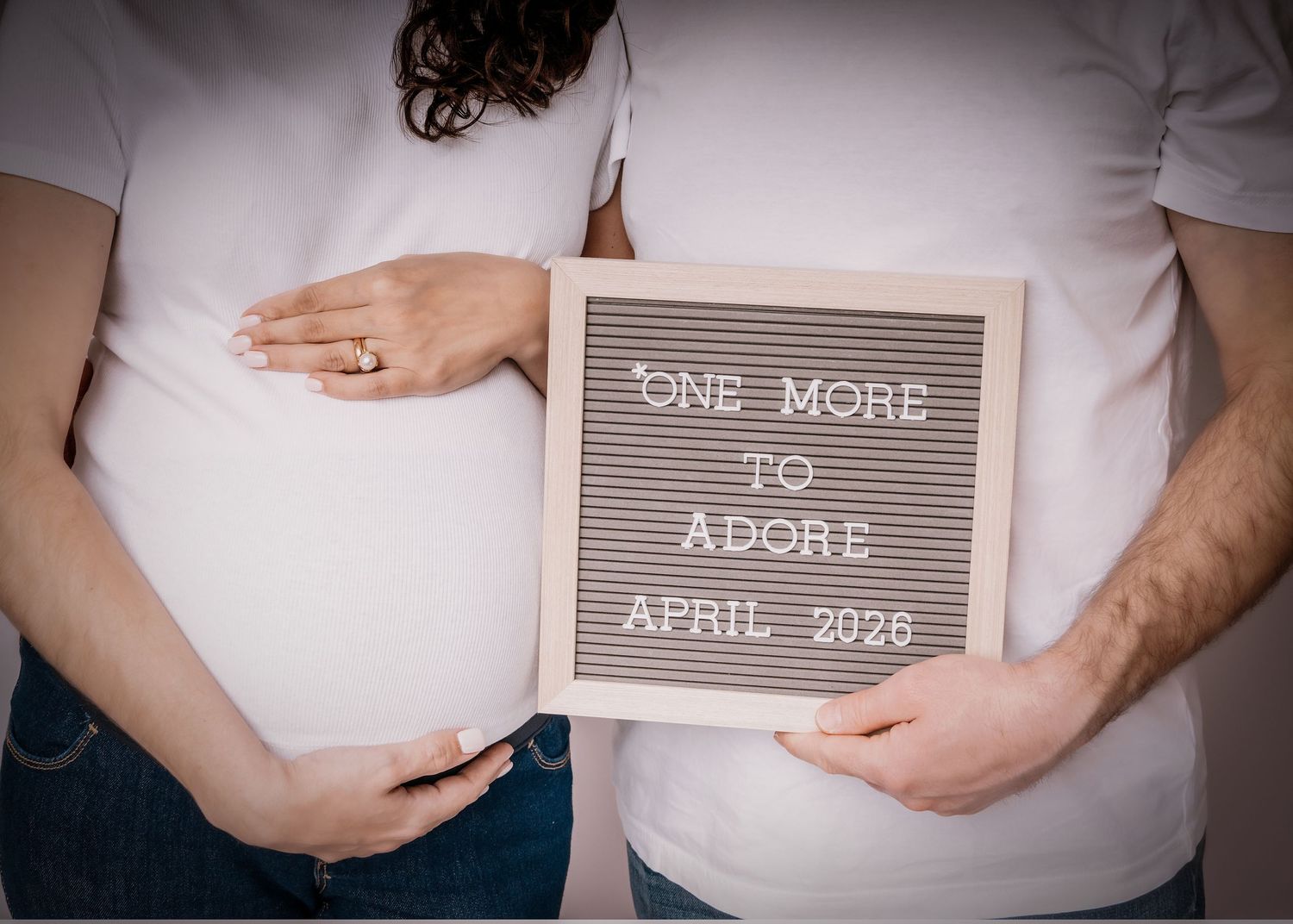 Pregnancy announcement photo with couple holding a letterboard sign reading One More To Adore April 2026.