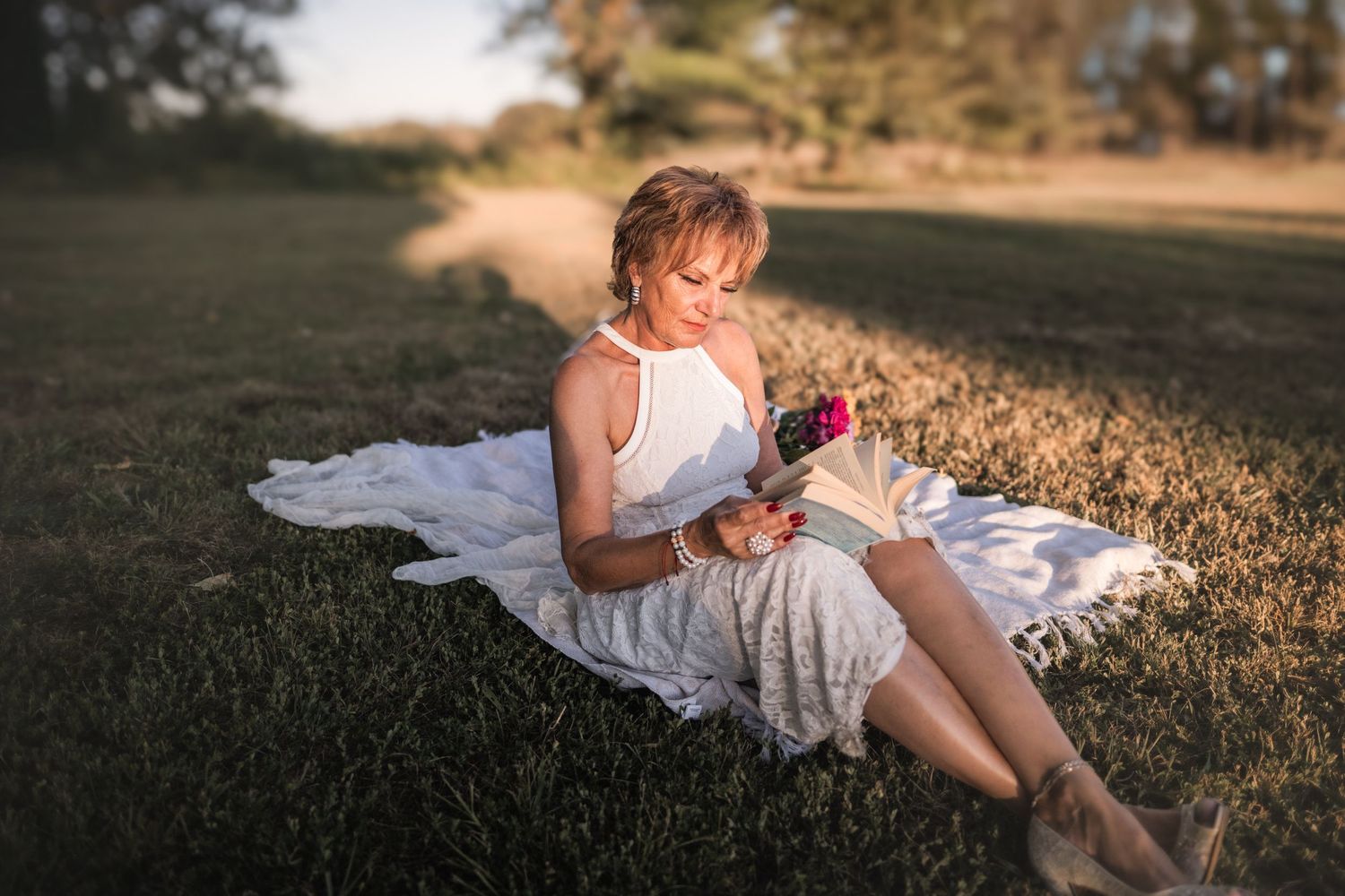 A peaceful outdoor scene with someone in white lace dress holding flowers on a blanket at sunset.