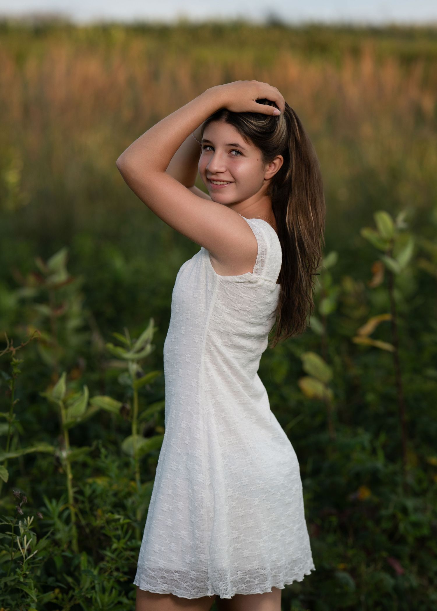Young girl in white lace dress poses in a lush green field, smiling over her shoulder with hand in hair.