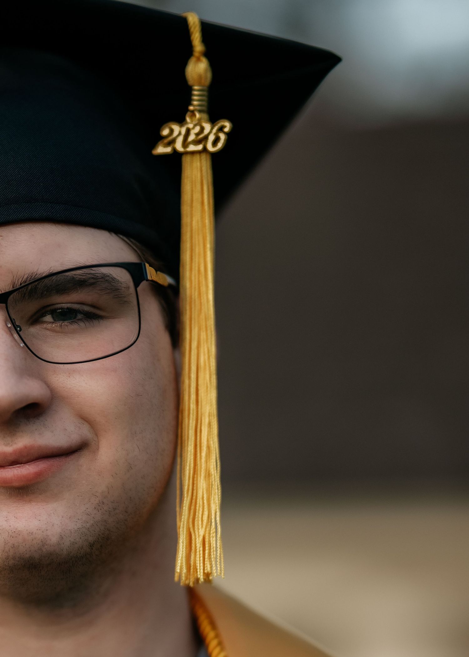 A young man wearing glasses and a black graduation cap with a gold 2026 tassel smiles proudly.
