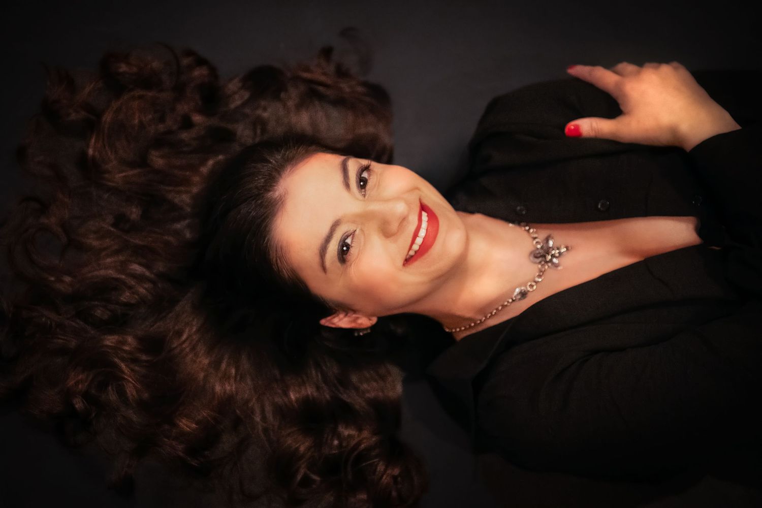 A smiling woman with curly dark hair lying down, wearing a black blazer and silver necklace, with red lips.