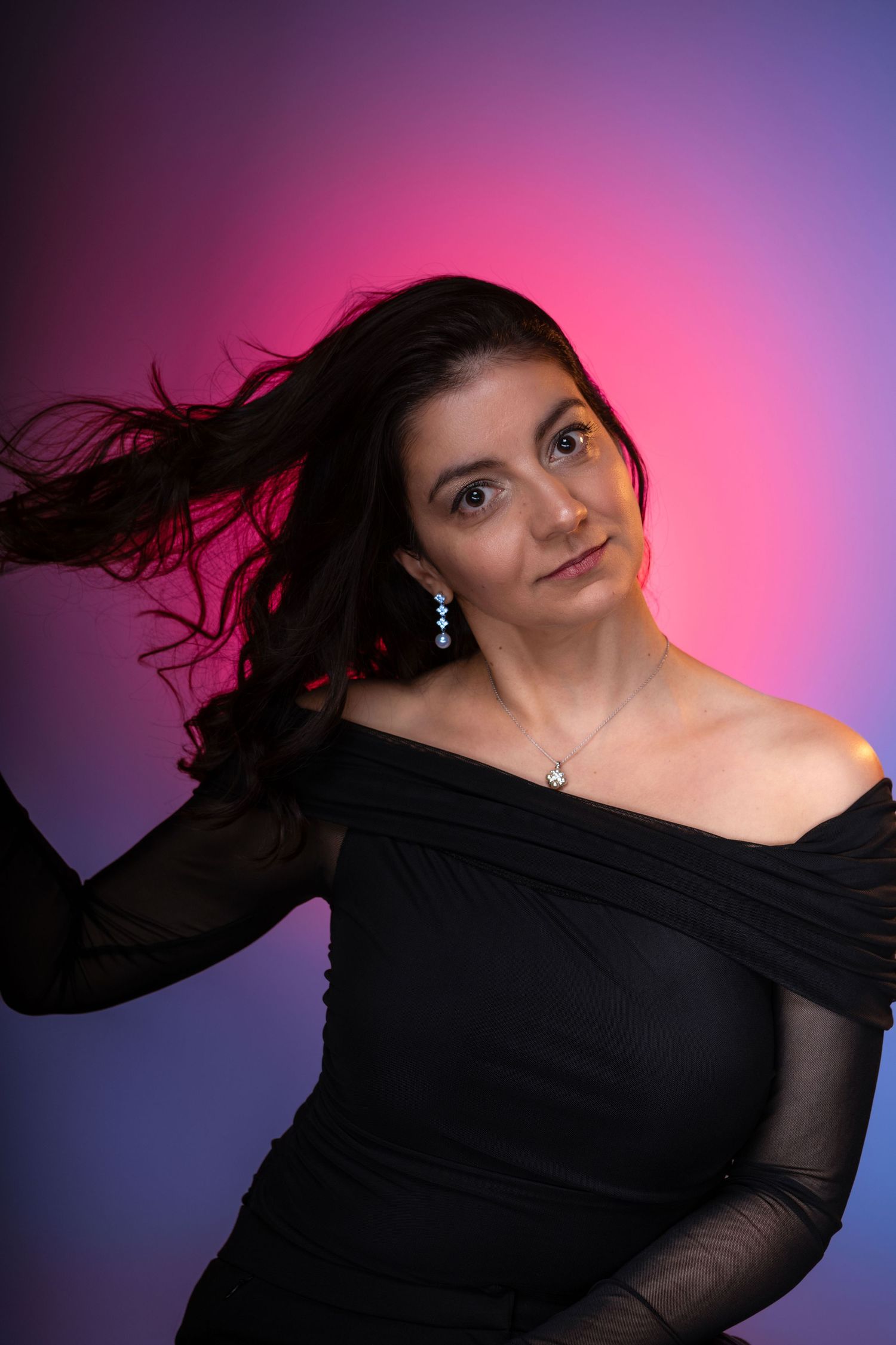 Woman in black off-shoulder dress with flowing dark hair poses against vibrant pink and purple gradient background.