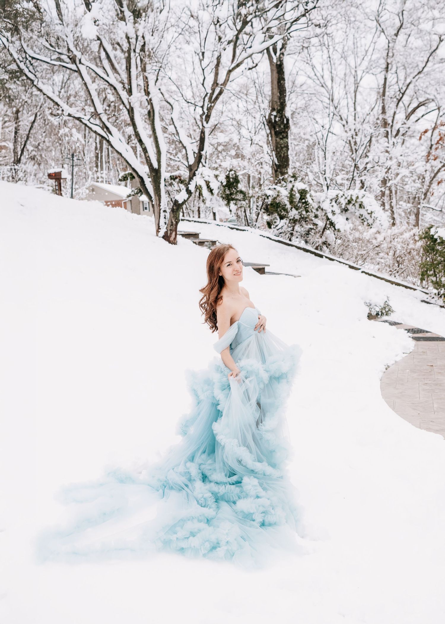 Woman in a flowing light blue tulle gown poses gracefully in a snowy winter landscape with snow-covered trees.