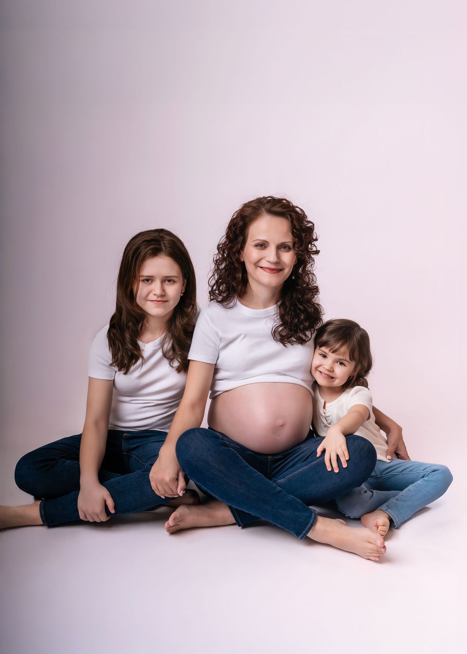 Pregnant woman in white top and jeans sits smiling with two young daughters in a bright studio maternity photo.