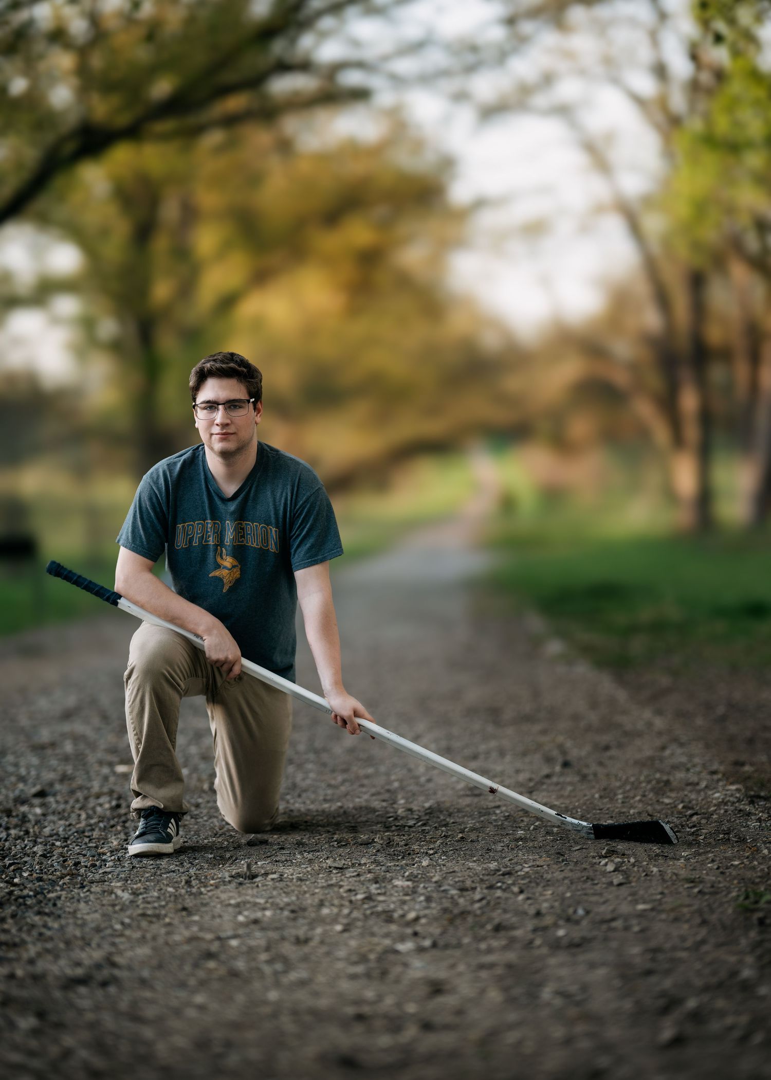 Young man in glasses kneeling on a gravel path holding a hockey stick, wearing a teal Vikings shirt.