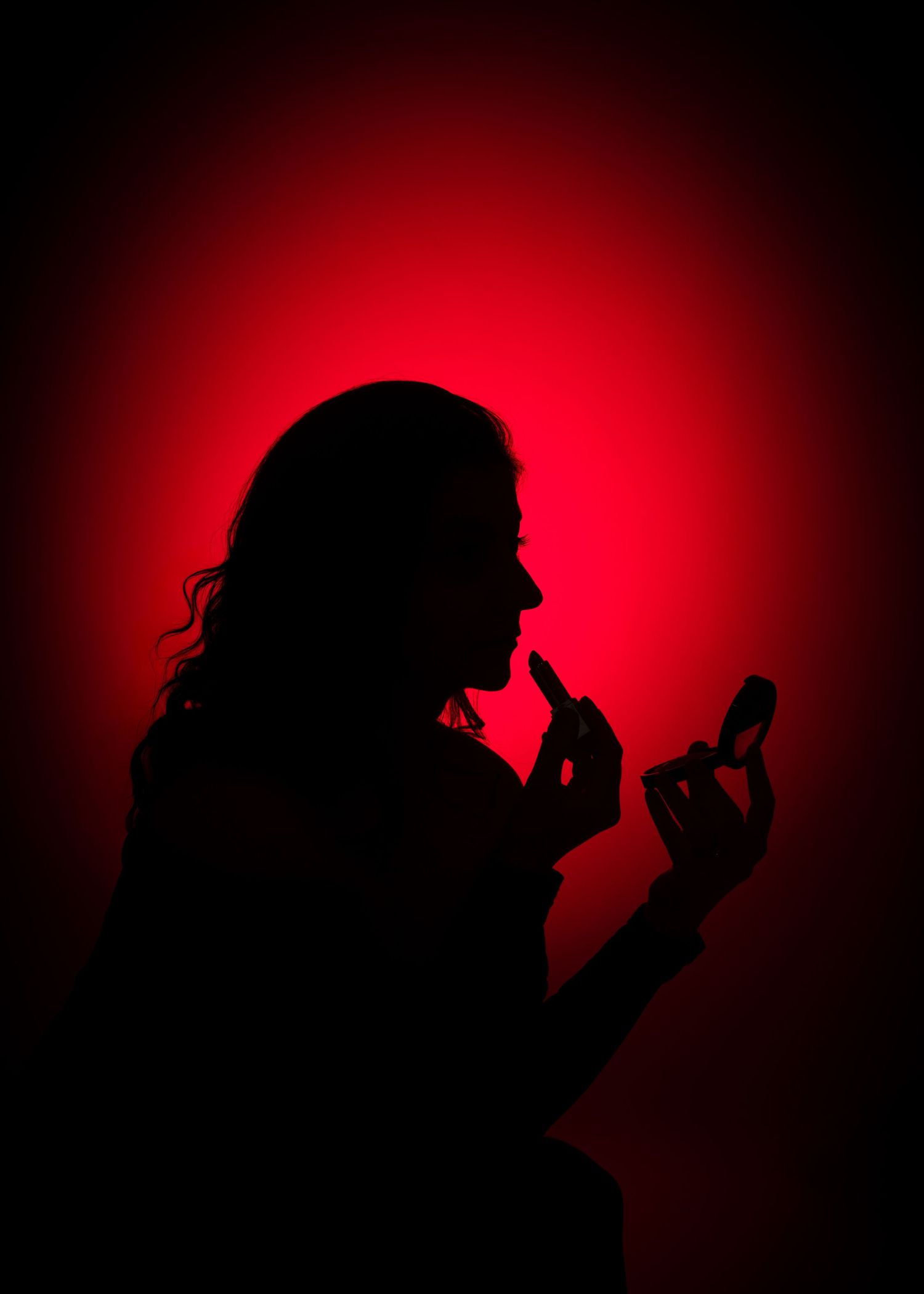 Silhouette of a woman applying lipstick against a dramatic red glowing background.