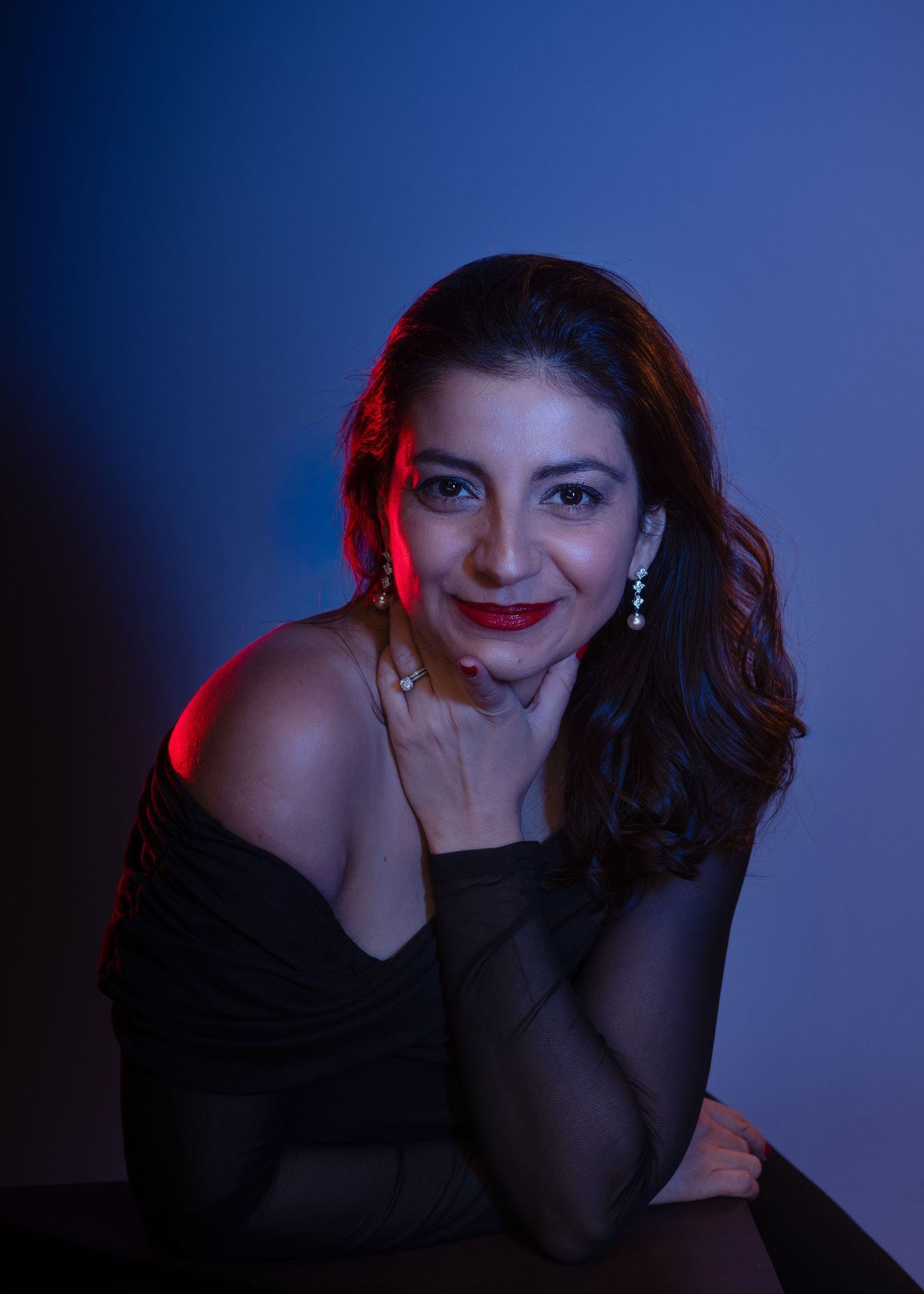 Woman in black off-shoulder dress poses elegantly under dramatic blue and red studio lighting with diamond earrings.
