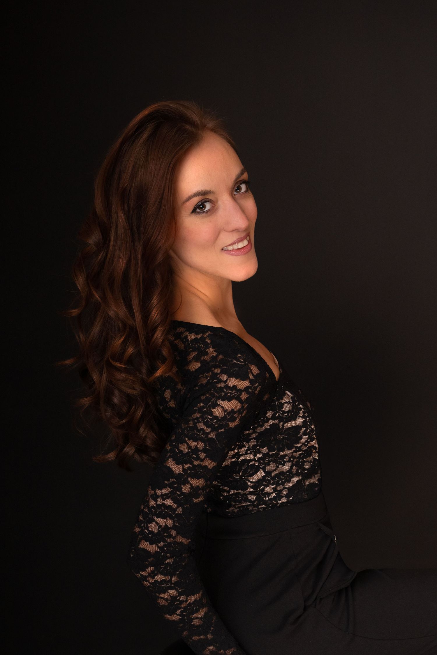 A smiling woman with curly auburn hair poses in a black lace dress against a dark studio background.