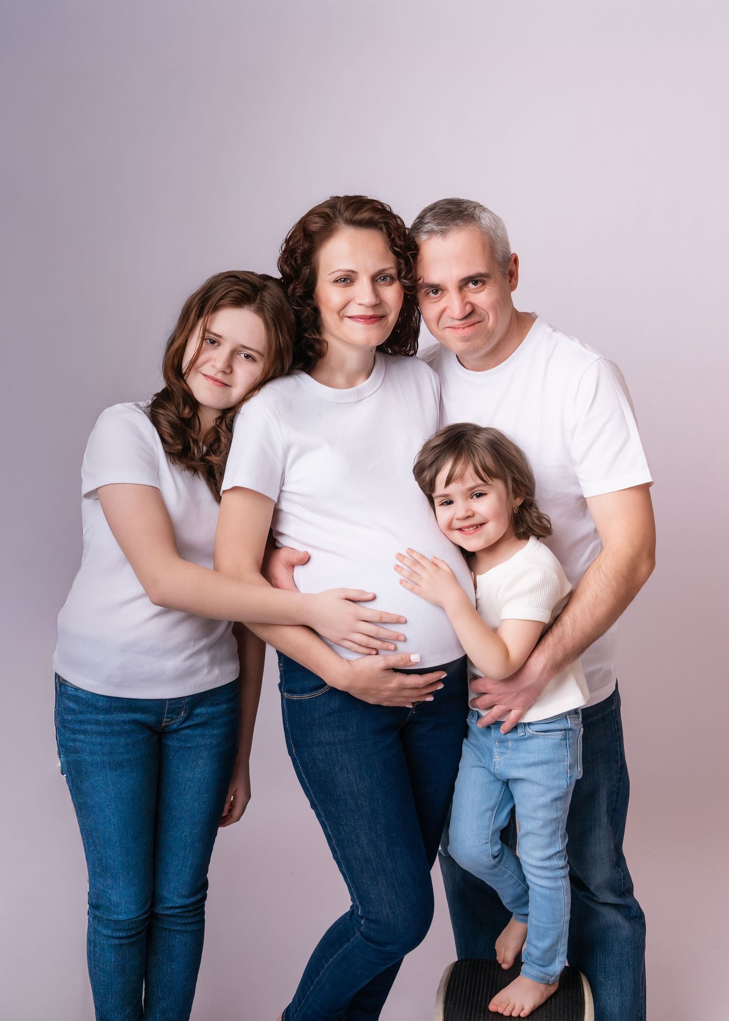 A pregnant woman poses with her family in matching white shirts and jeans against a light background.