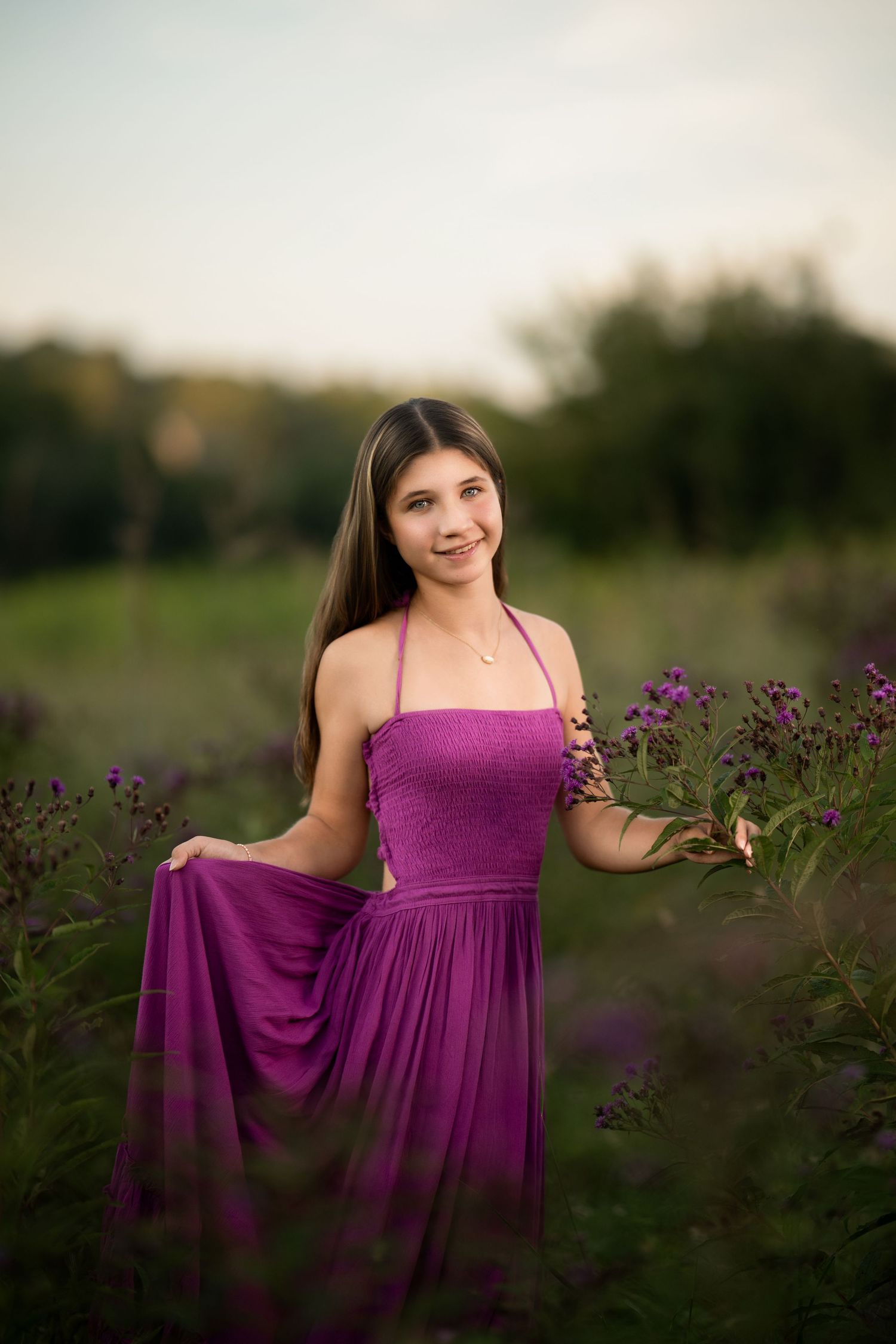 Young woman in purple halter dress smiling among wildflowers in a lush green field at golden hour.