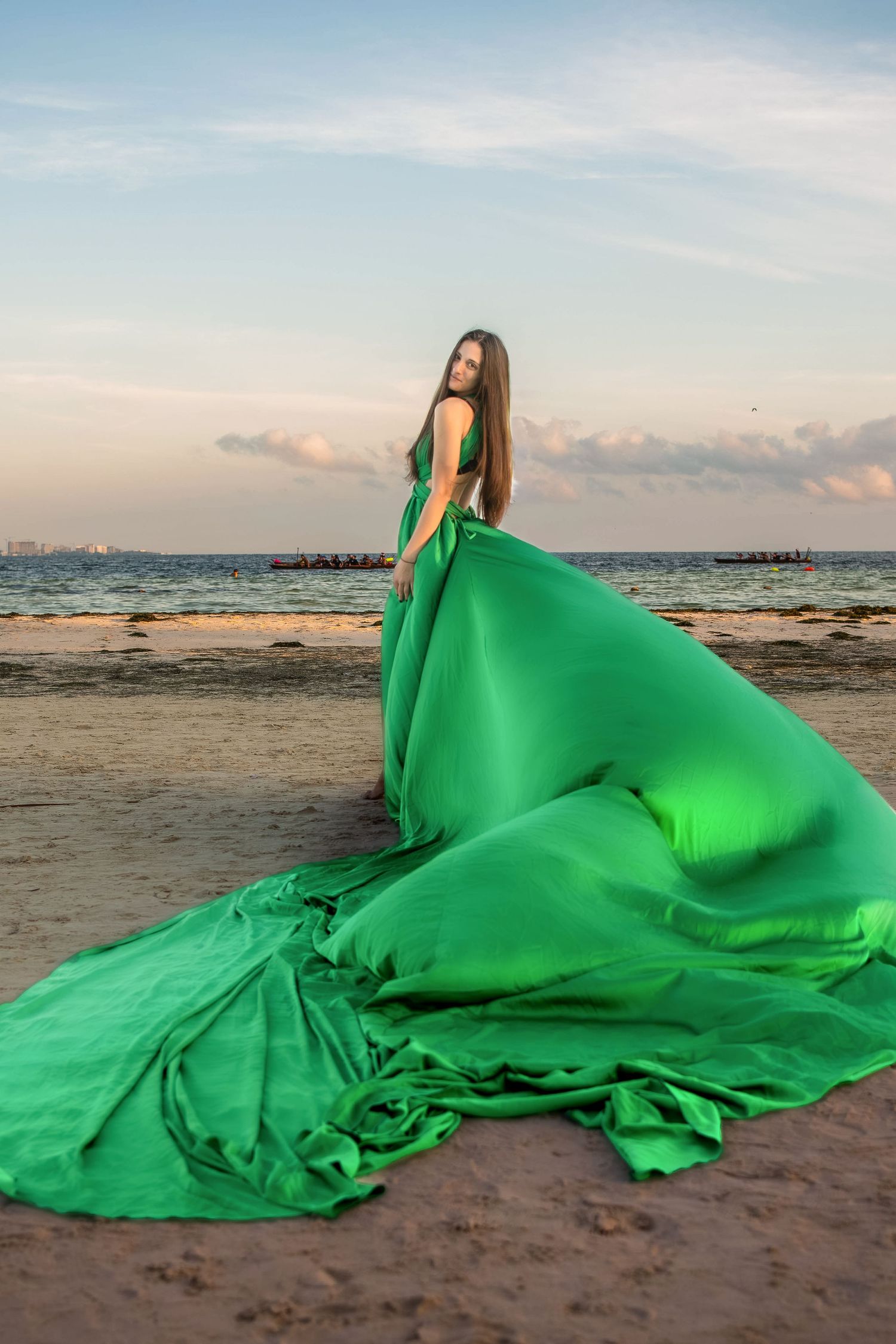 Woman in flowing green gown poses on sandy beach at sunset with ocean waves in background.
