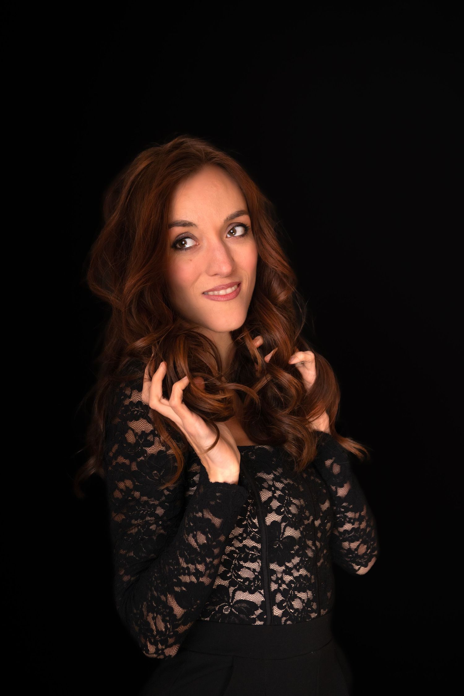 A young woman with long auburn wavy hair poses in a black lace dress against a dark background.