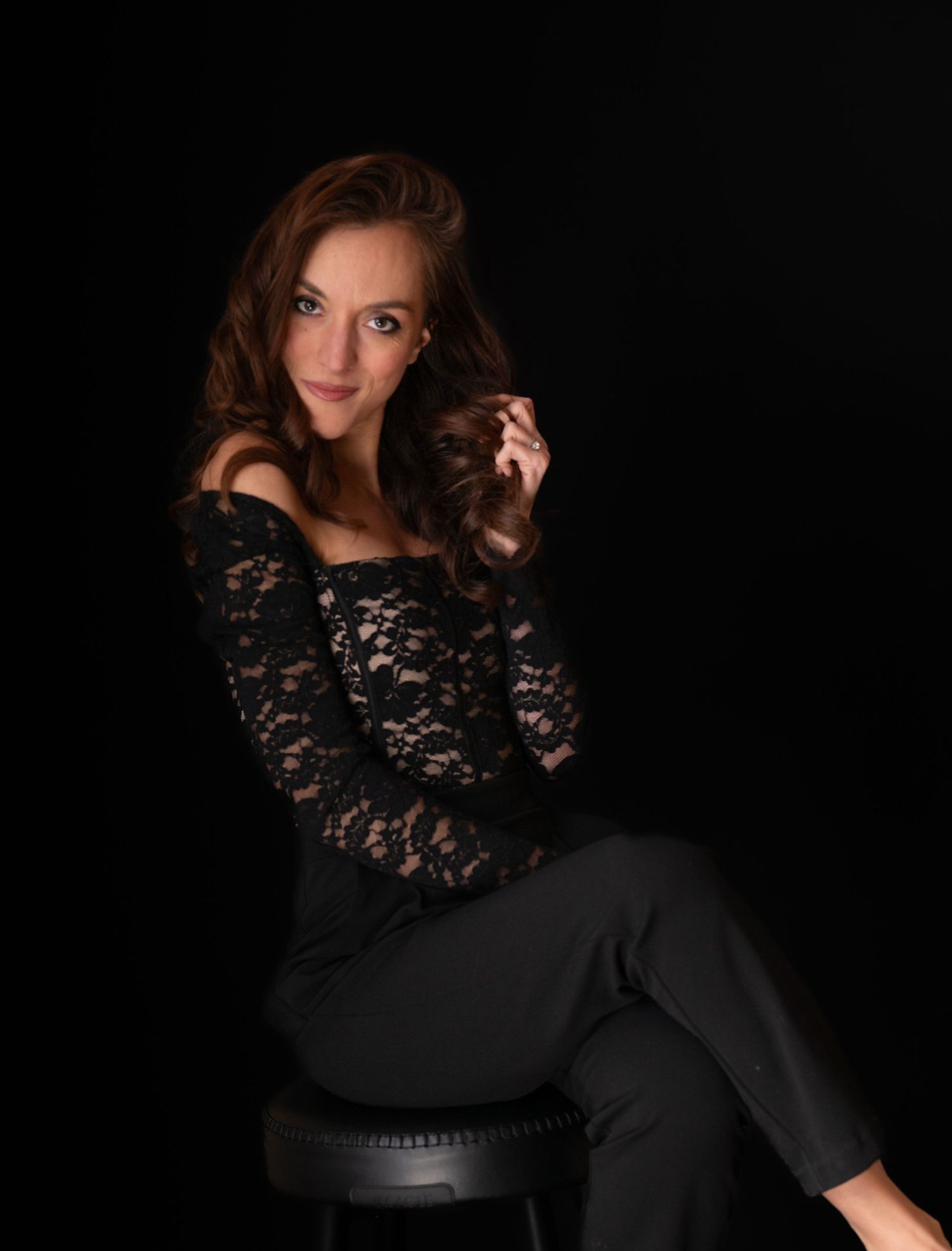 Woman in black lace top and dark pants poses elegantly on a stool against a dark studio background.