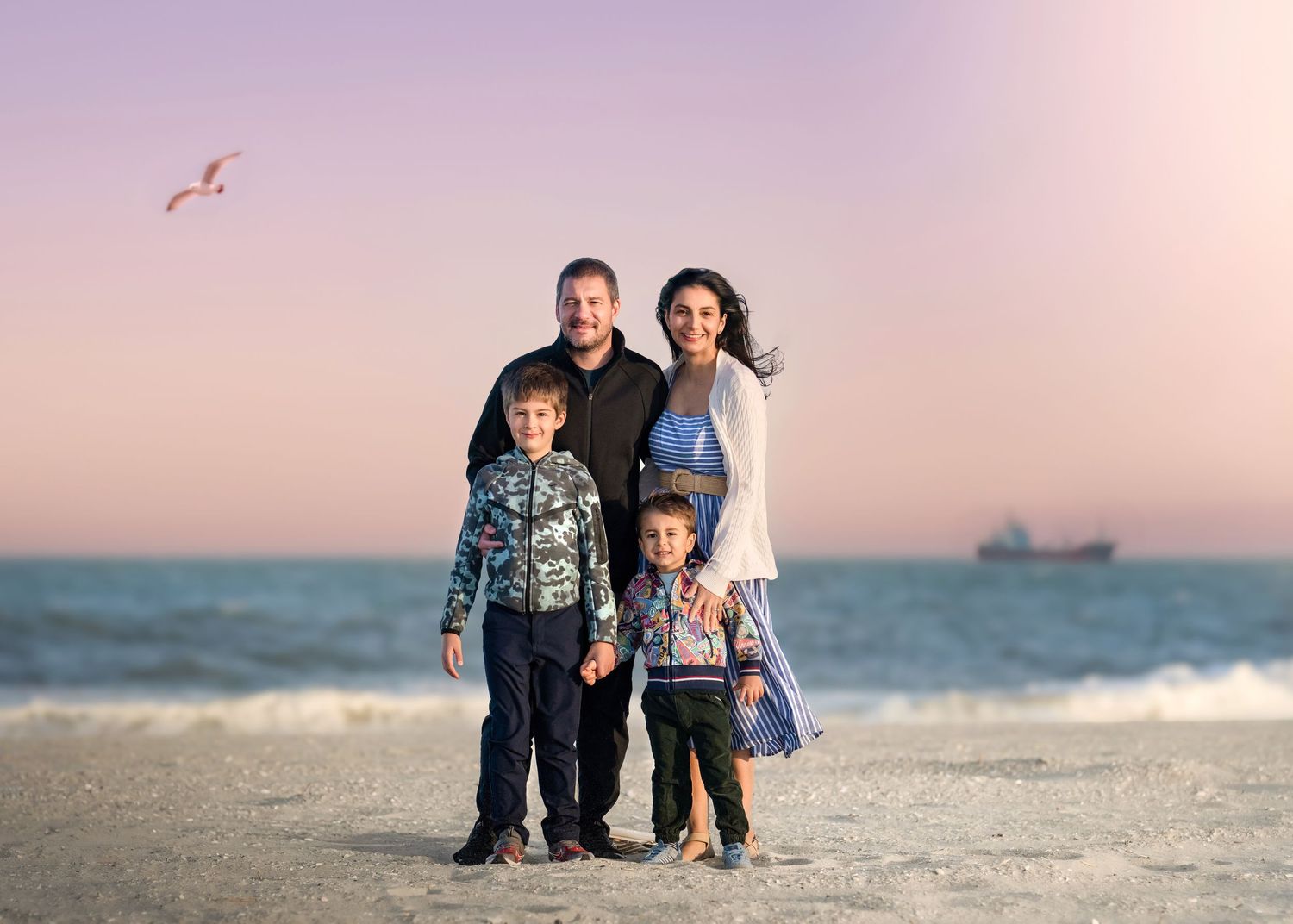 Family of four posing on a sandy beach at sunset with ocean waves and a ship in the background.