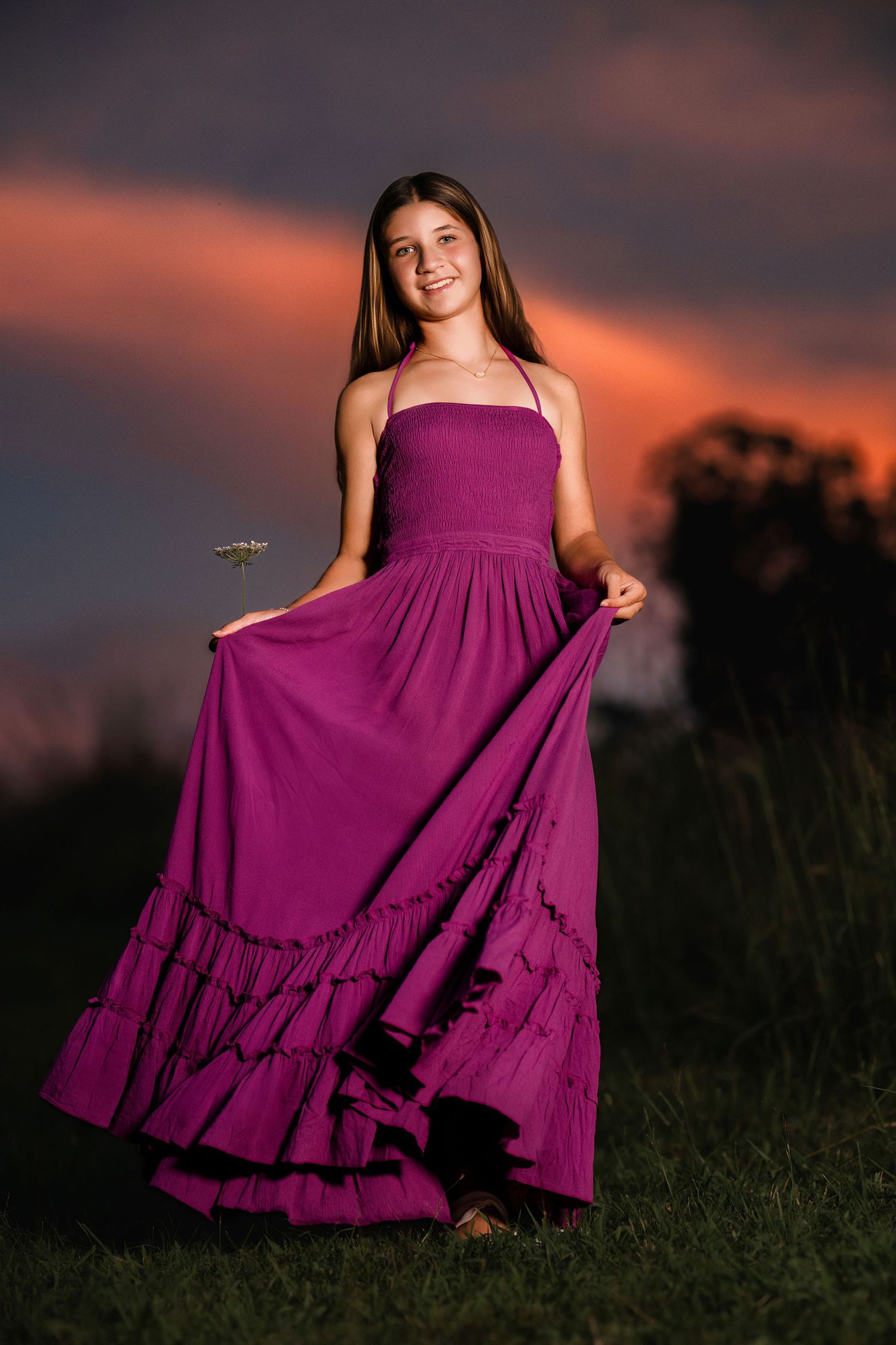Teen girl in purple maxi dress twirling at sunset with dramatic pink and orange sky backdrop.