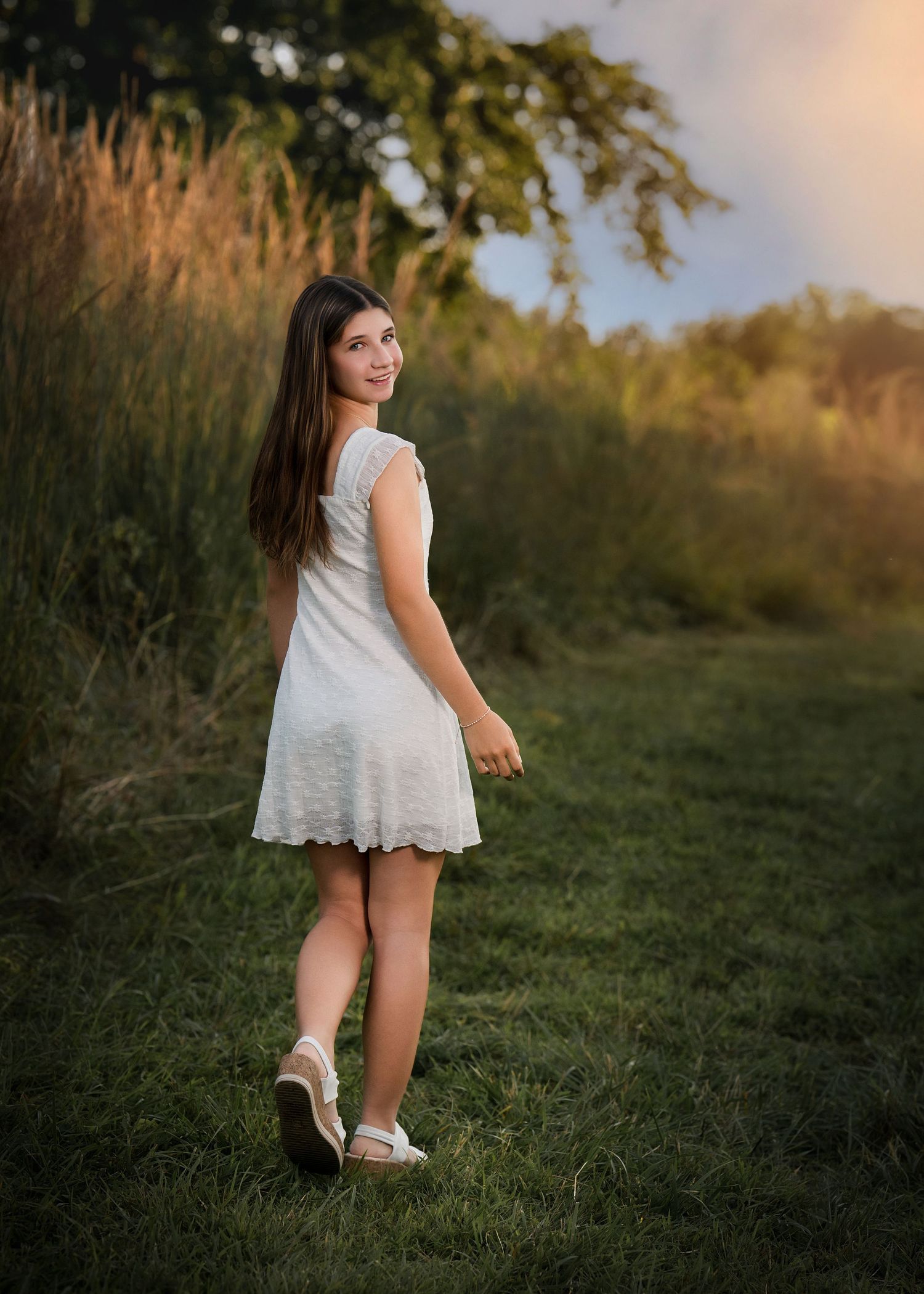 A young girl in a white dress walks through a lush green field at golden hour, glancing back over her shoulder.