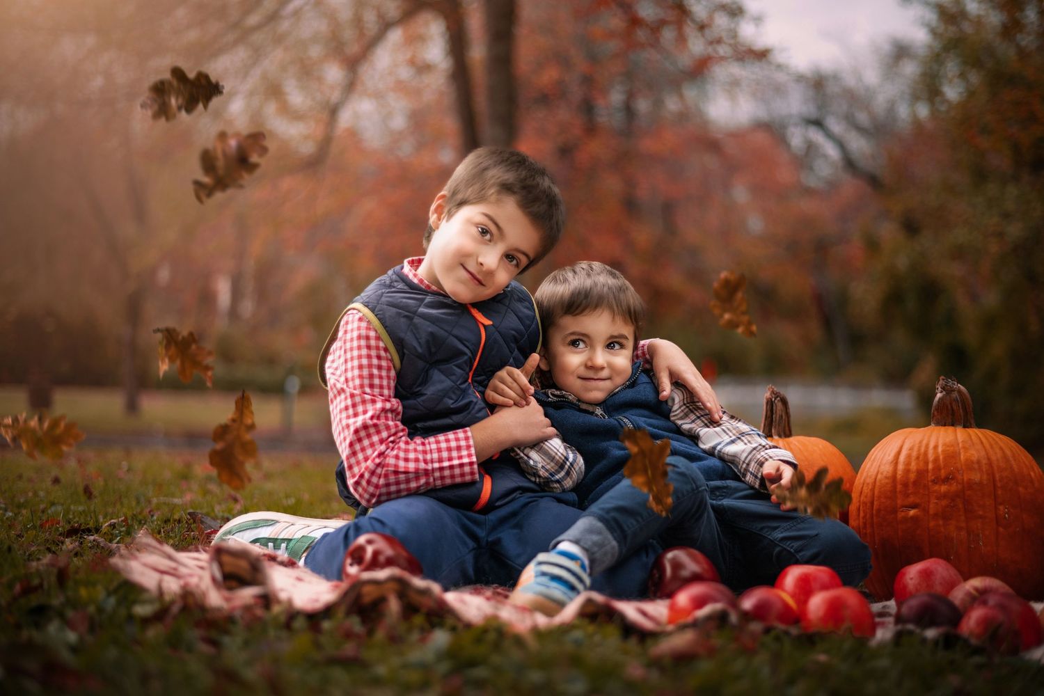 Two young boys hugging on a blanket in an autumn park with pumpkins and apples during fall foliage season.