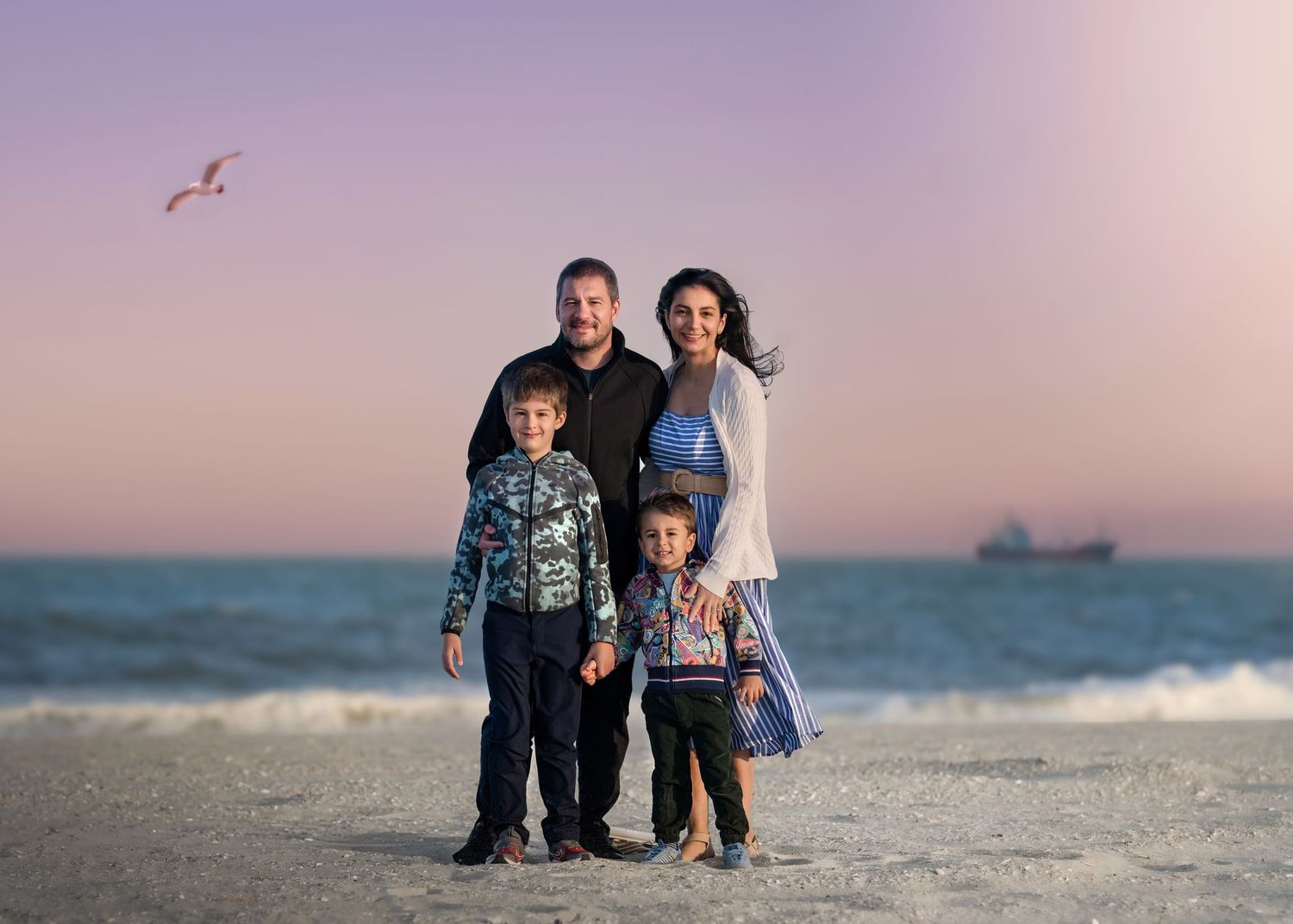 Family of four posing on a beach at sunset, with ocean waves, a ship, and a bird in the pastel sky behind them.