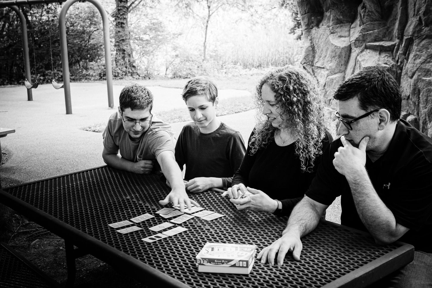 Four friends playing cards at an outdoor metal table in a park, black and white photography.