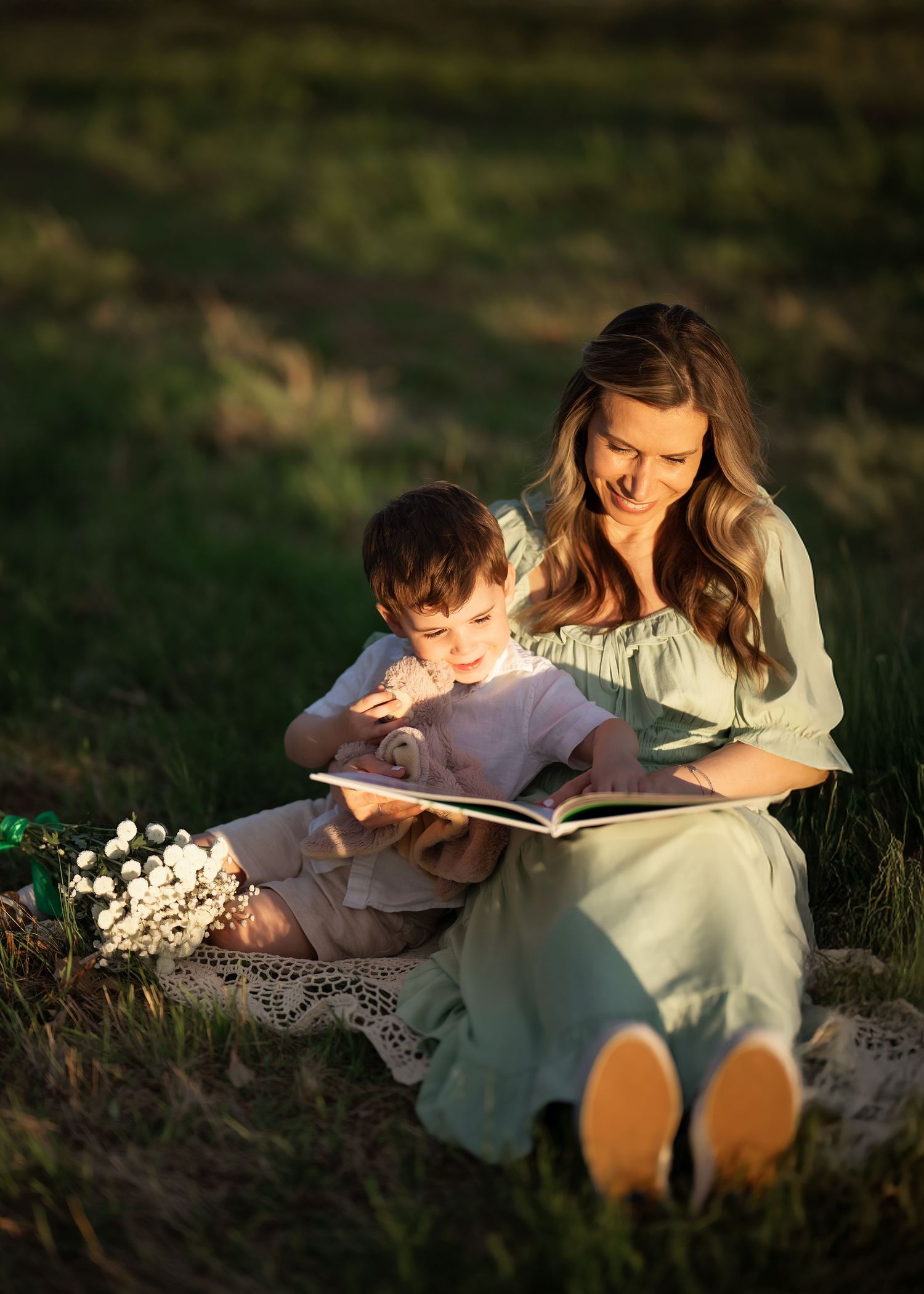 A smiling mother in a green dress reads a book with her young son outdoors on a blanket in golden sunlight.