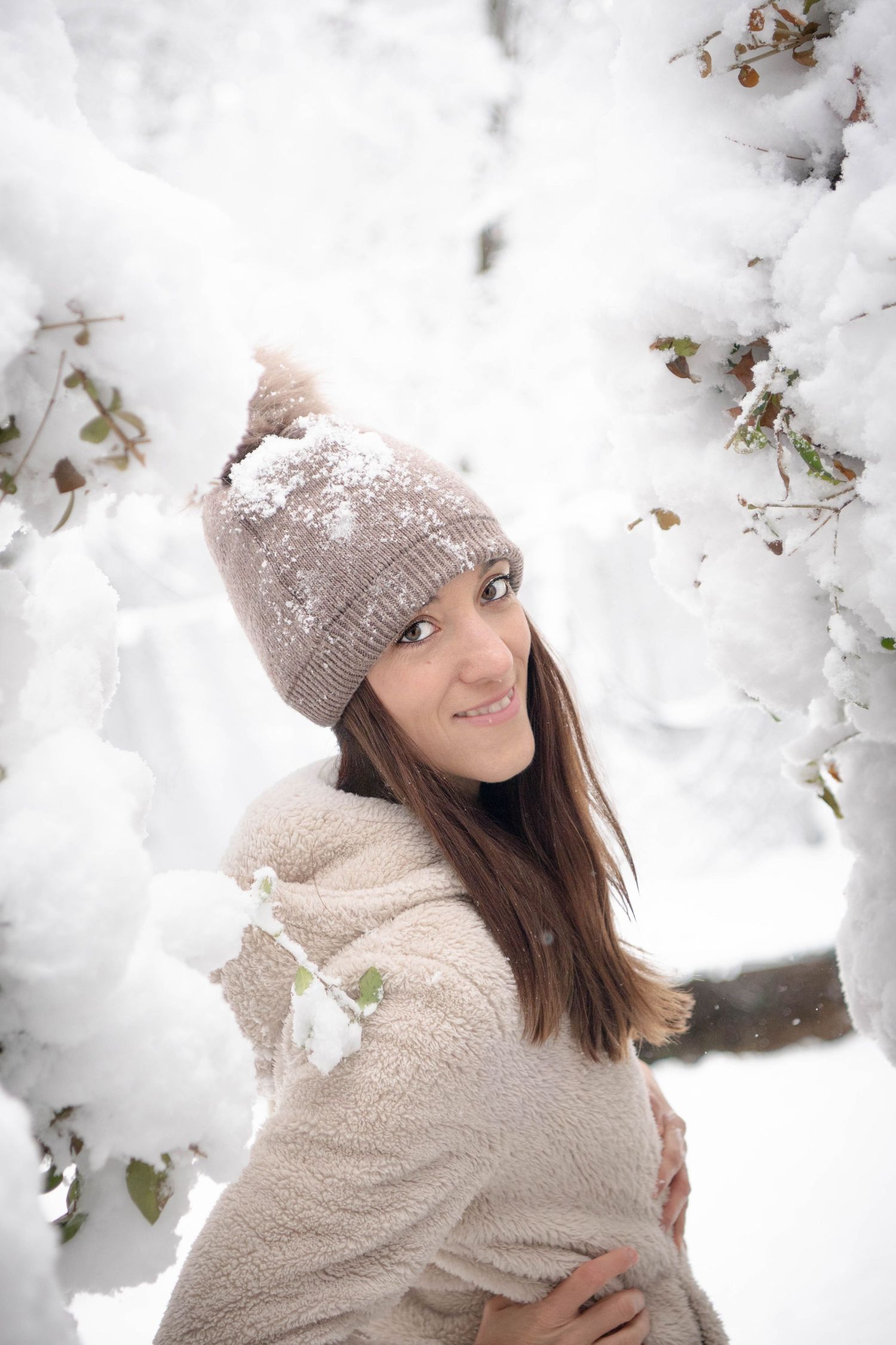 Person wearing beige winter clothing poses among snowy tree branches.
