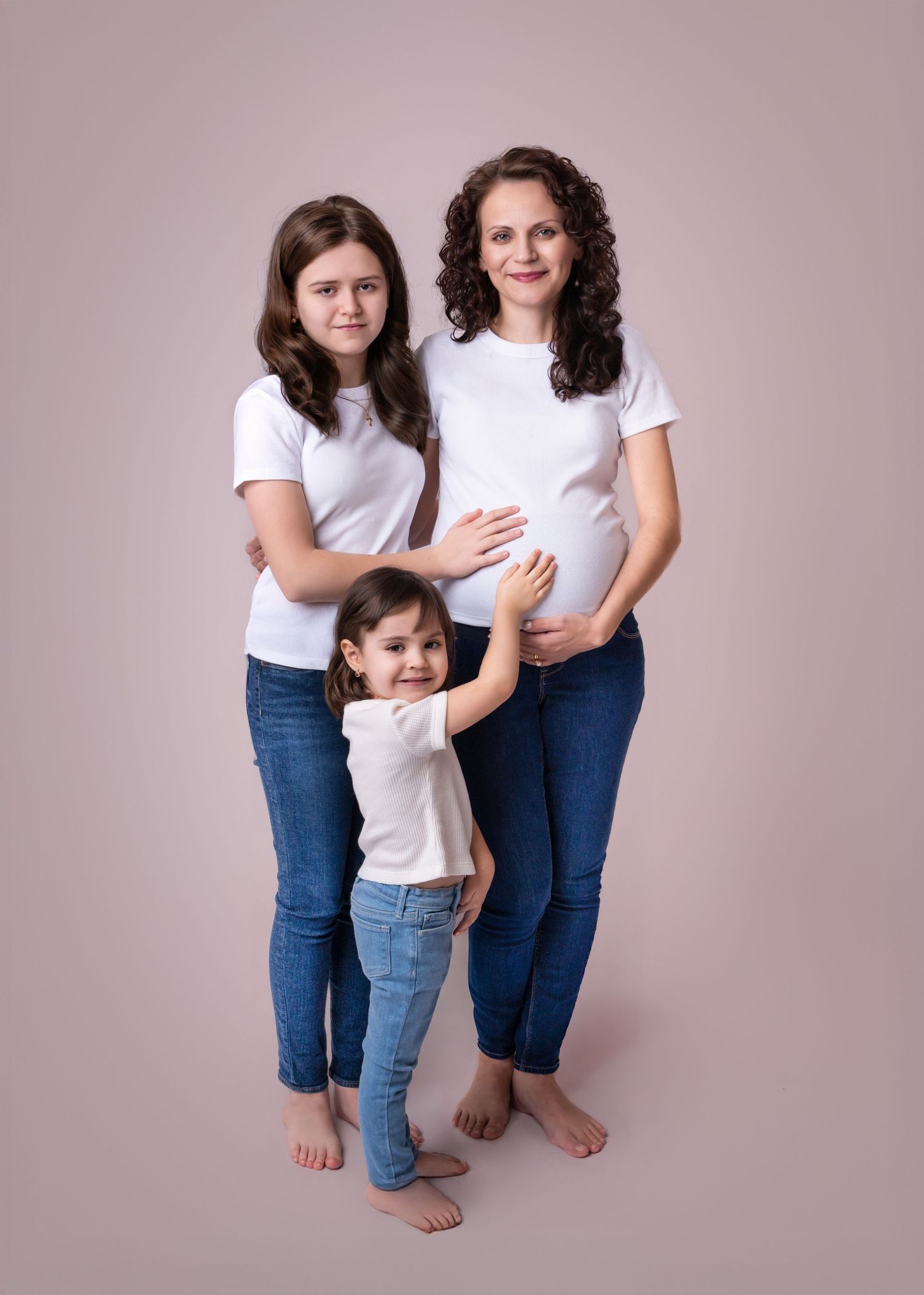Pregnant mother poses with teen daughter and toddler girl, all in white shirts and jeans, barefoot.