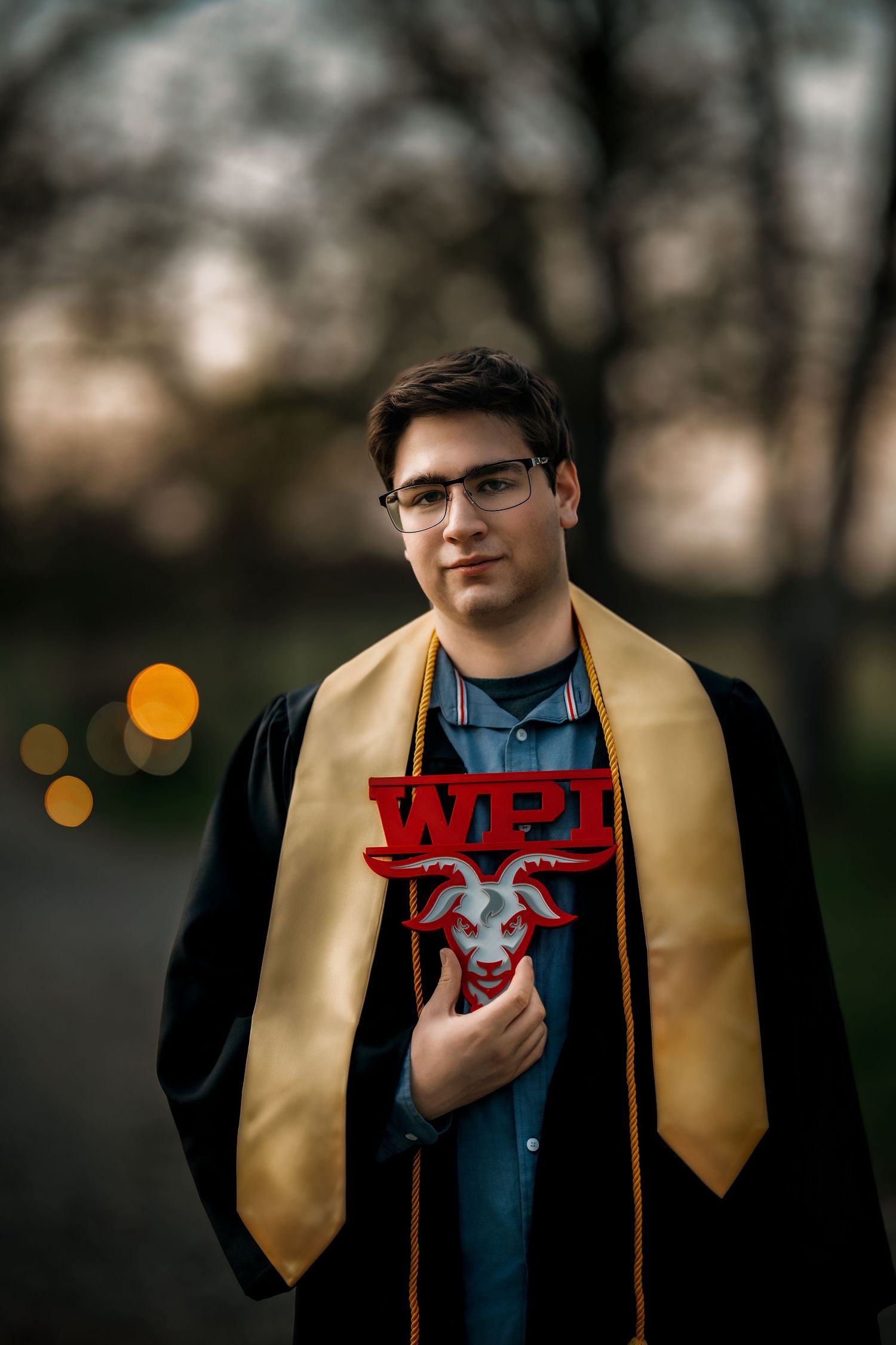 WPI graduate in black gown and gold stole holds red WPI logo sign with goat mascot at dusk.