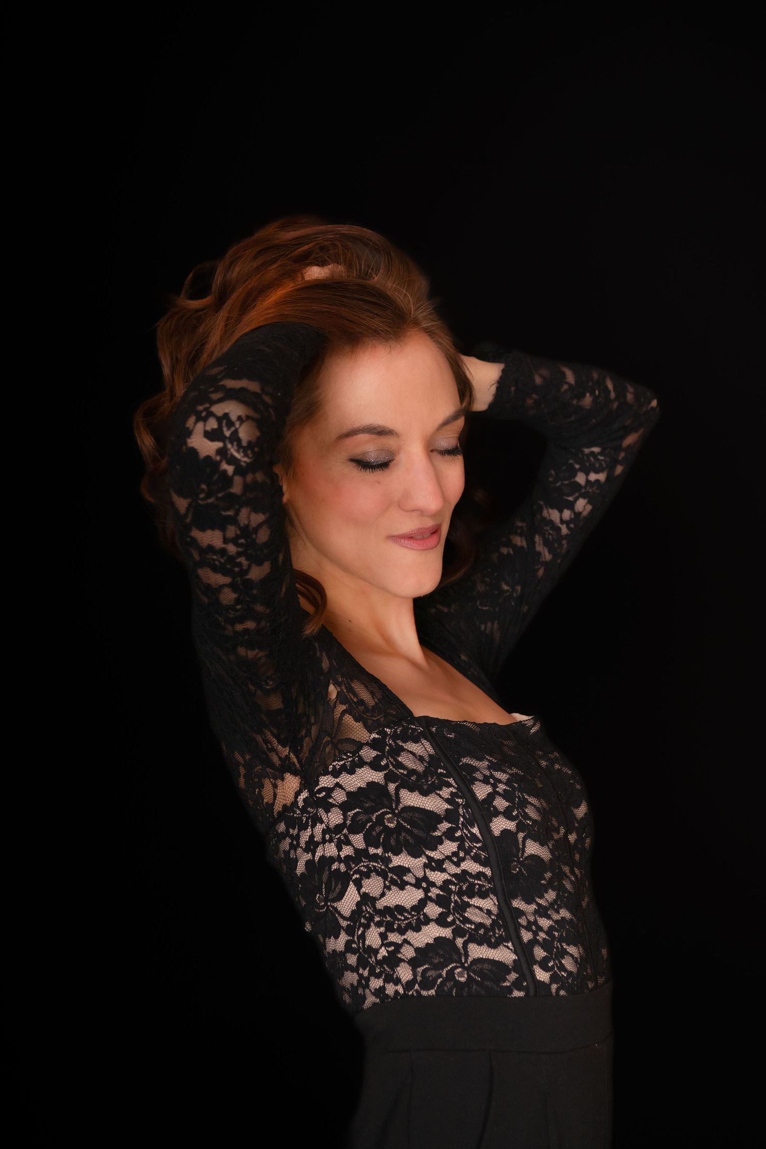 Woman in black lace top poses with hands in auburn hair against dark background.