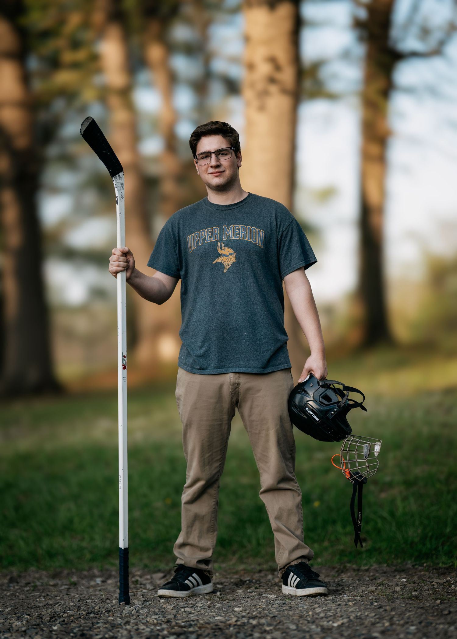 Young man in Upper Merion shirt holds hockey stick and helmet outdoors among trees.