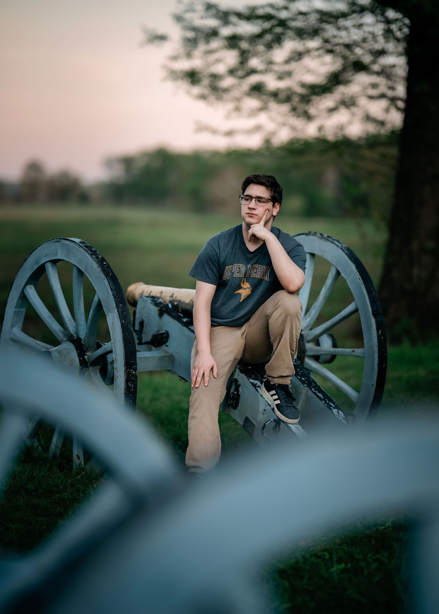 Young man in glasses sits on a historic Civil War cannon at dusk in a green field at golden hour.