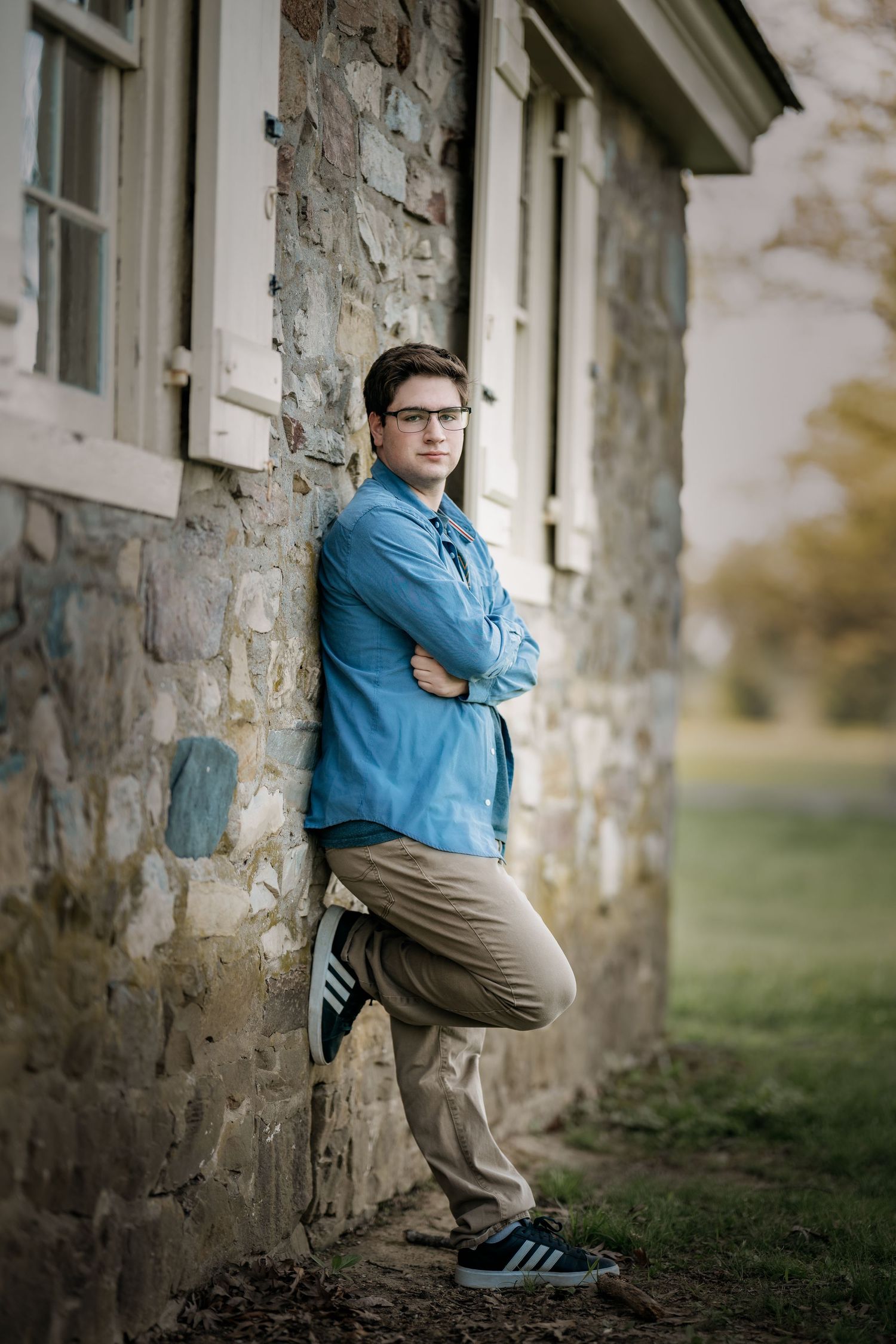 Young man in blue shirt and glasses leaning against stone building with arms crossed.