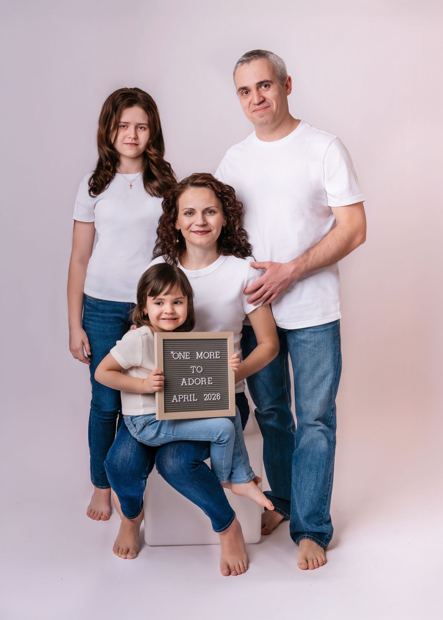 Family of four in white tees and jeans holding a sign reading One More to Adore April 2025, announcing a pregnancy.