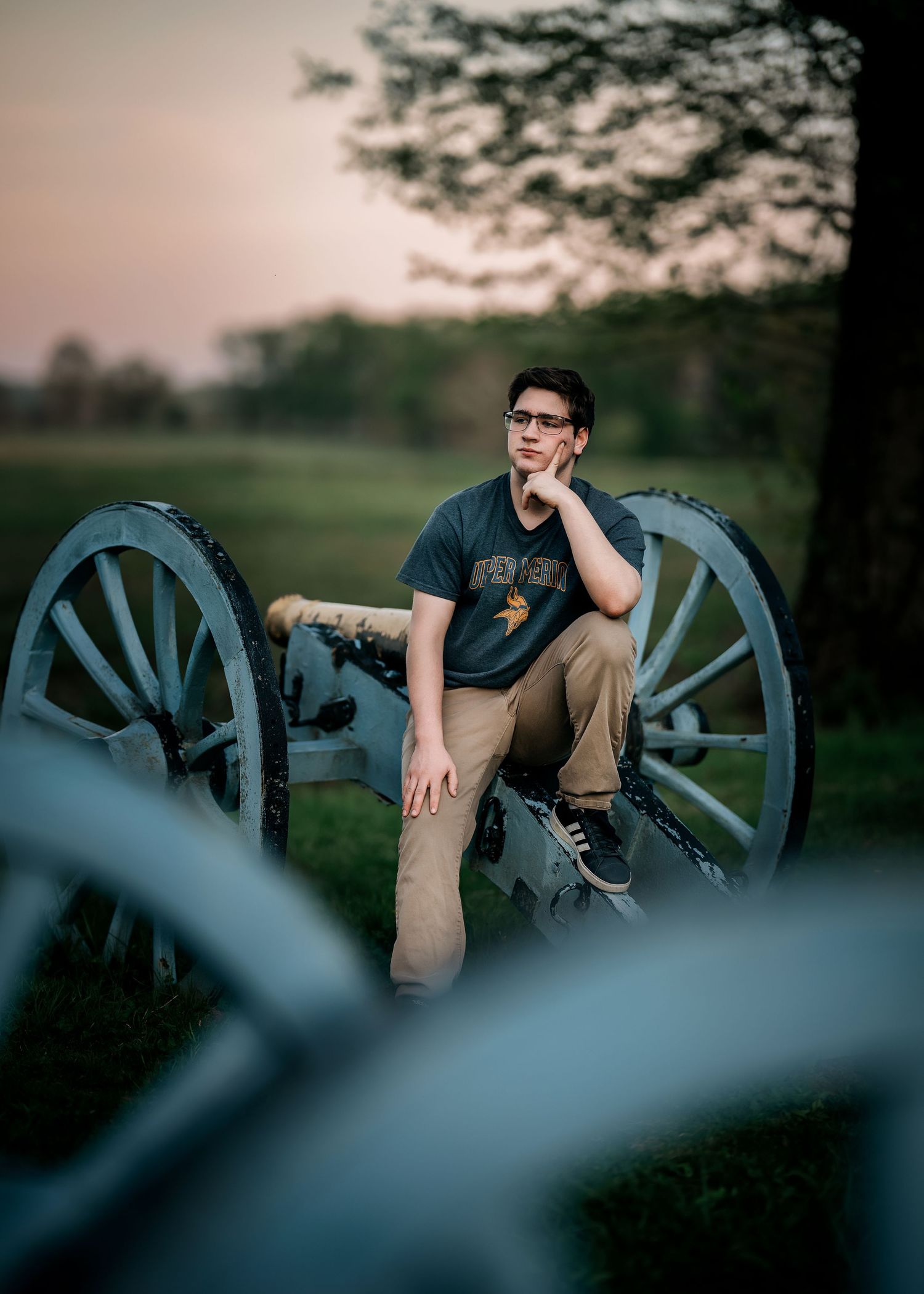 Young man in glasses sits on a historic Civil War cannon at dusk in a grassy battlefield setting.