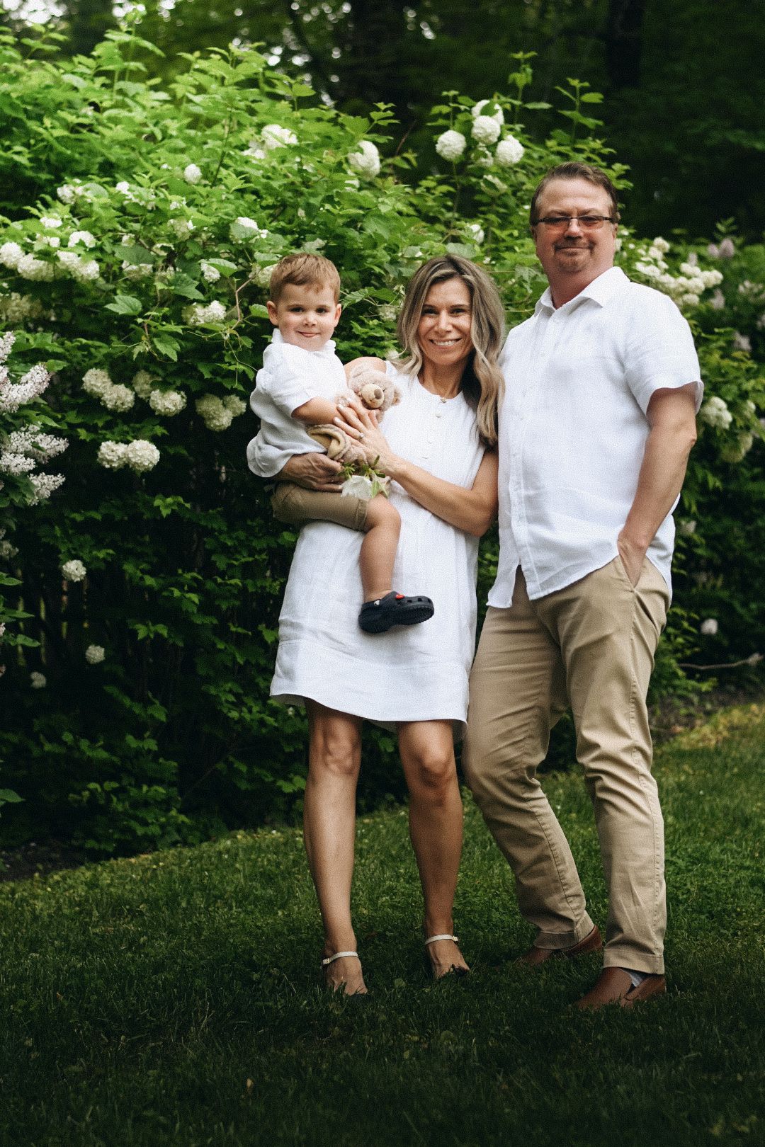 Family of three posing outdoors in white outfits near blooming hydrangea bushes on a sunny day.