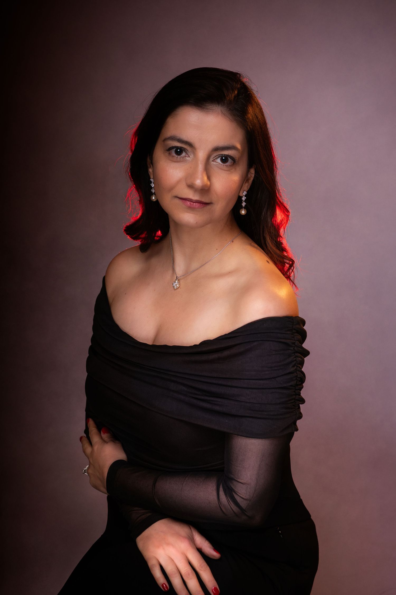 Woman in black off-shoulder dress with silver jewelry poses elegantly against a dark studio background with red lighting.