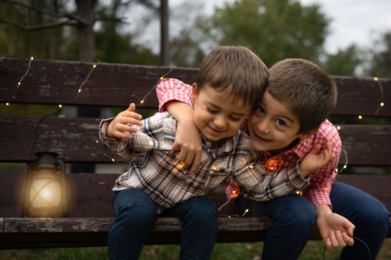 Two children in plaid and gingham shirts share a sweet moment on a rustic wooden bench at sunset.