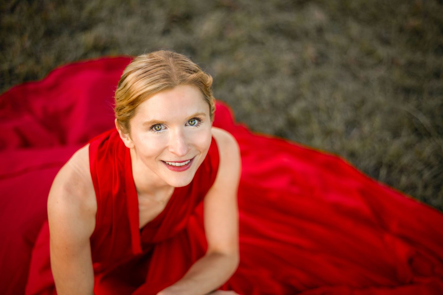 Woman in flowing red dress poses gracefully on grass during outdoor photoshoot.