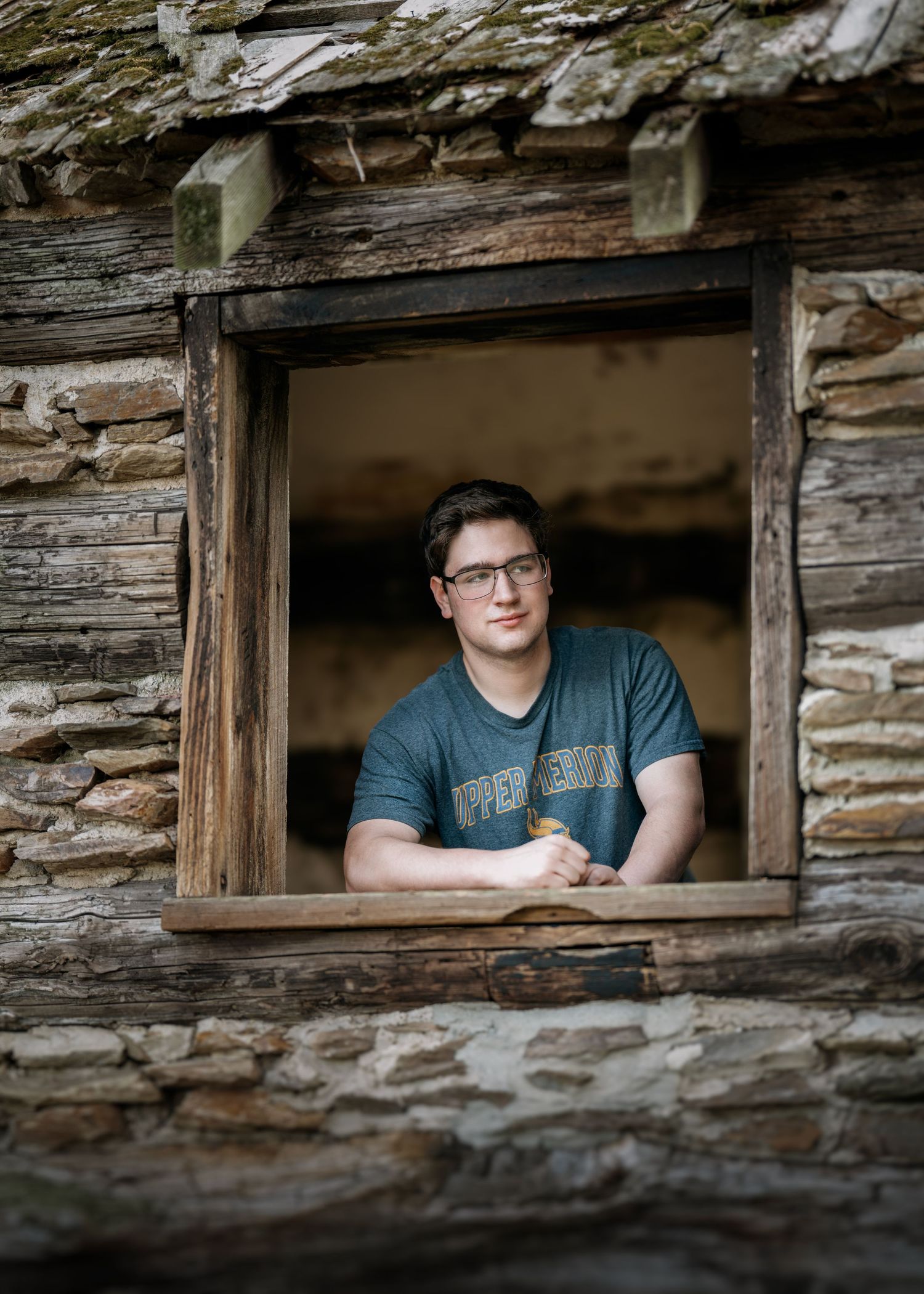 Young man with glasses leaning on rustic stone building window frame, wearing a teal graphic t-shirt.