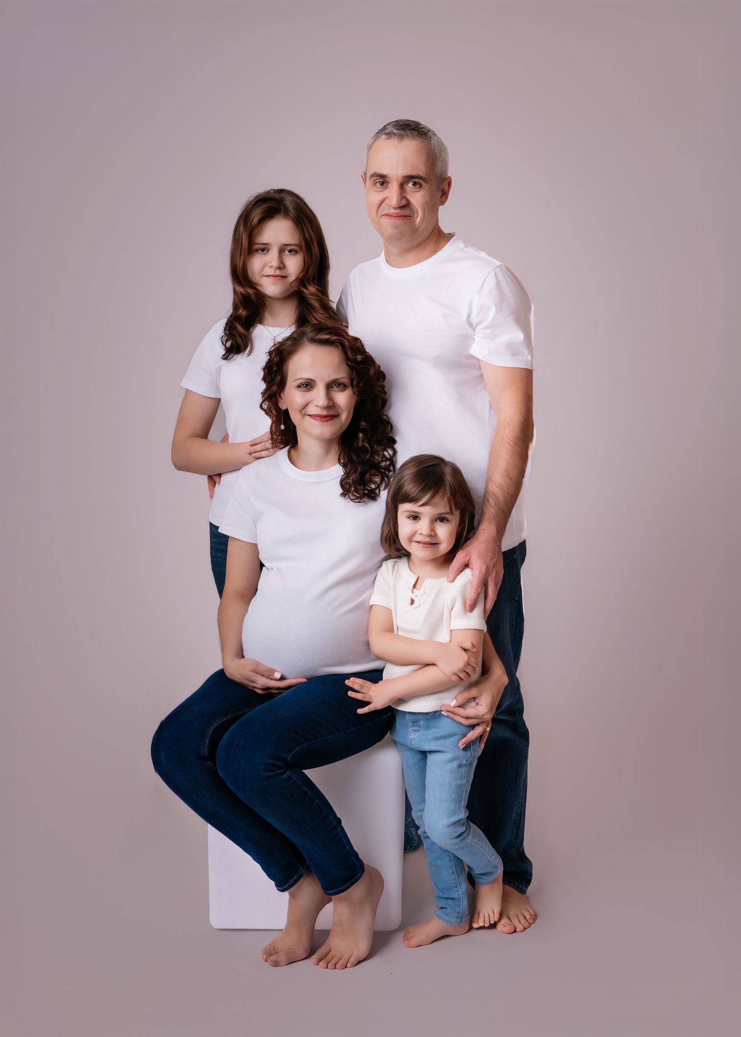 A happy family of four with a pregnant mother poses together in white shirts and jeans on a beige background.