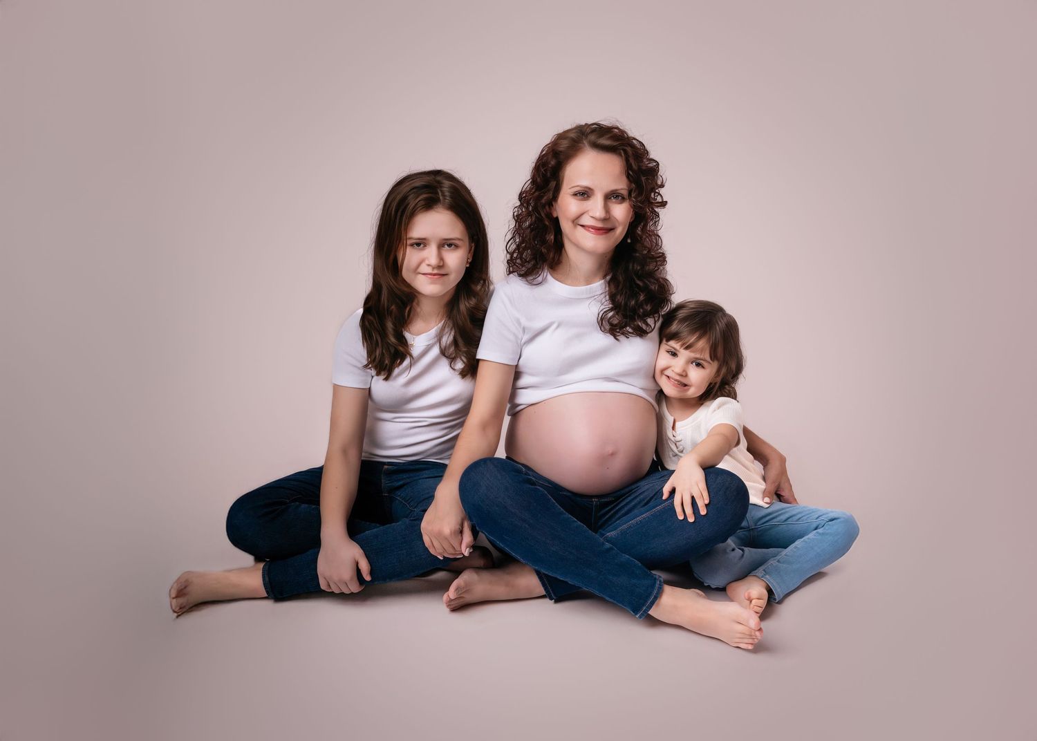 Pregnant woman in white top and jeans sits smiling with two young daughters on a neutral studio background.