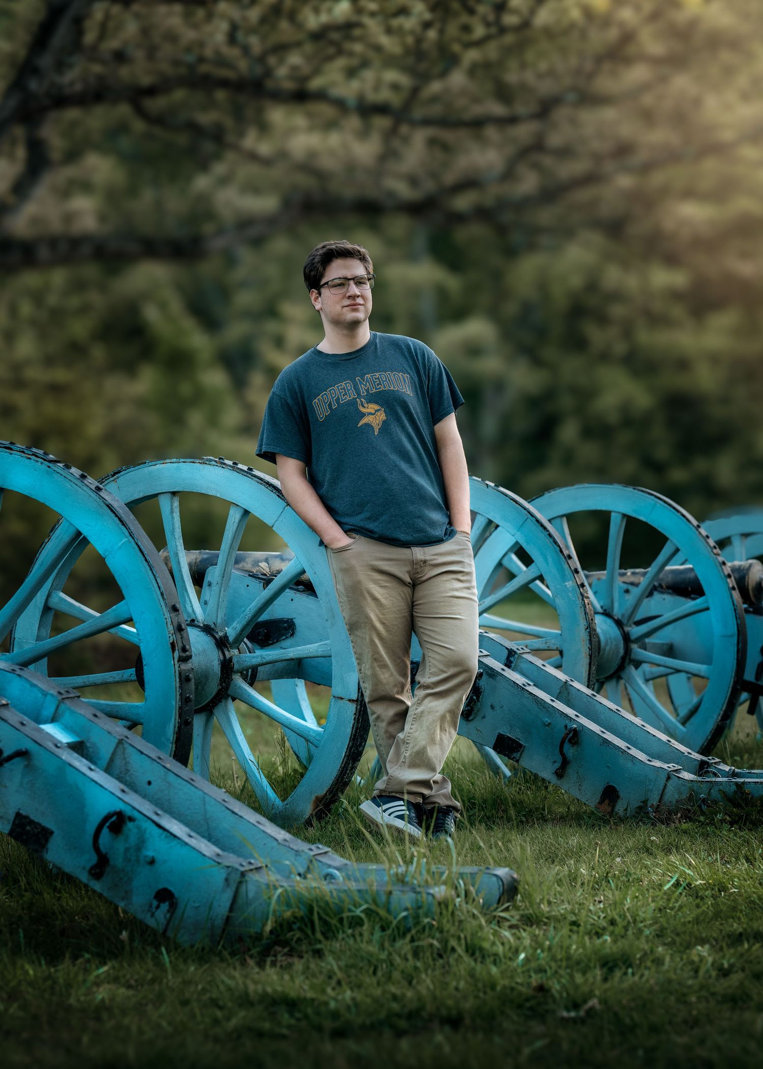 Young man in blue Vikings shirt poses confidently among vintage blue cannons on green grass at historical site.