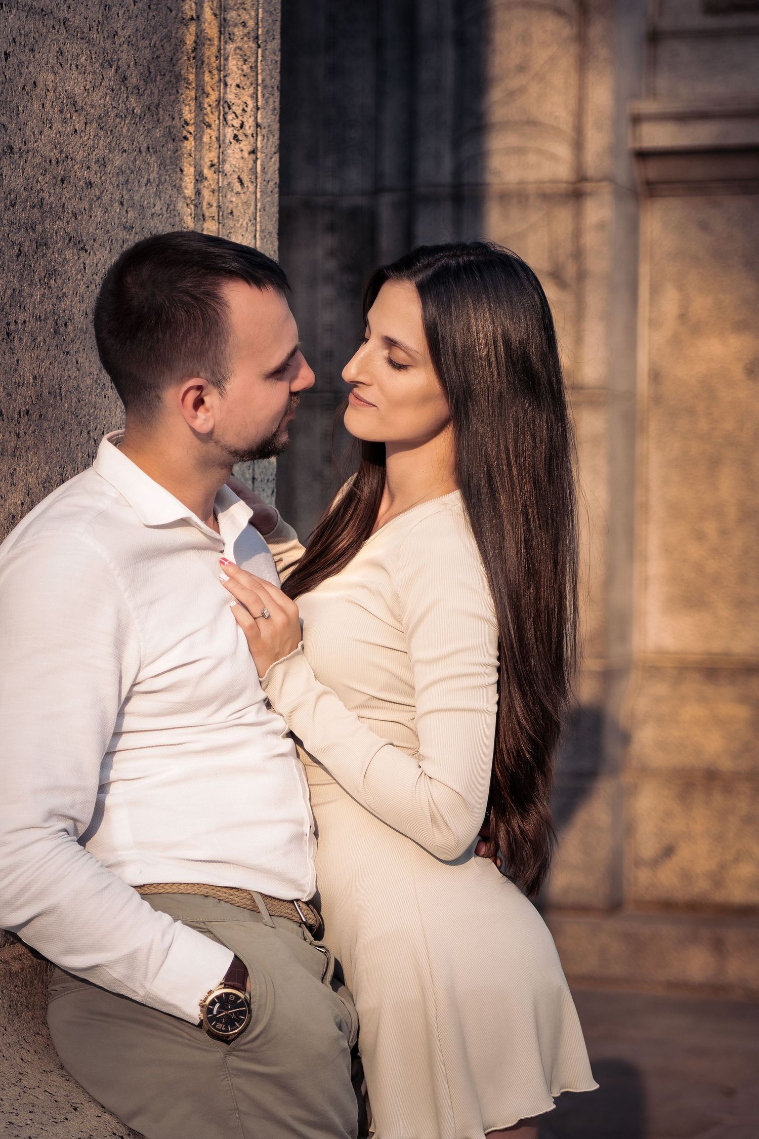 Romantic couple in elegant attire sharing an intimate moment against a stone column outdoors.