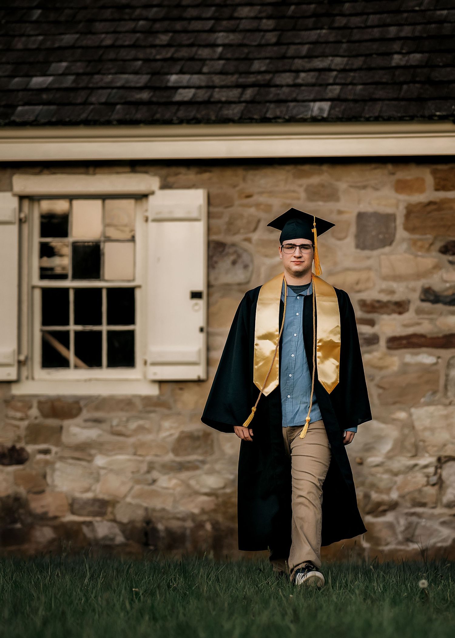 A young graduate walks proudly in black and gold cap and gown outside a historic stone building.