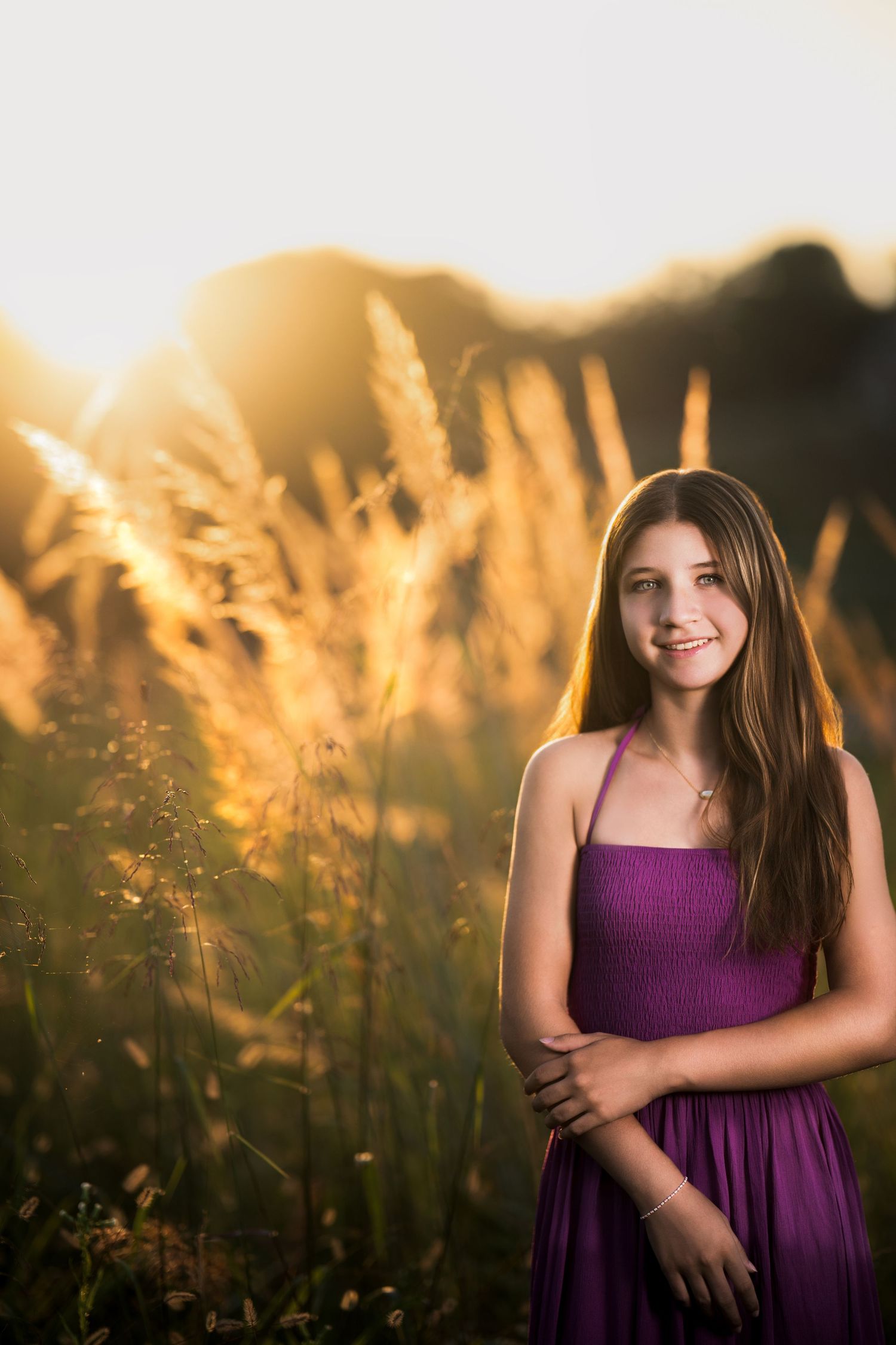 A smiling young girl in a purple dress poses in a golden sunlit field with tall grass during sunset.